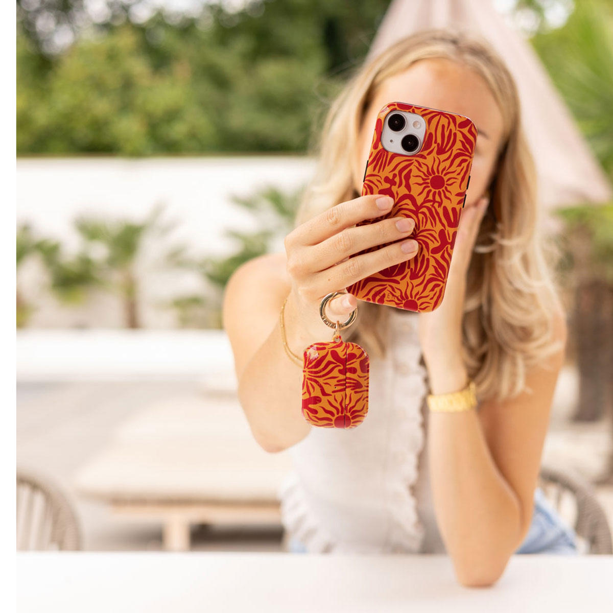 Blonde vrouw die een selfie maakt met een telefoonhoesje met patroon en een bijpassende AirPods-case buitenshuis.