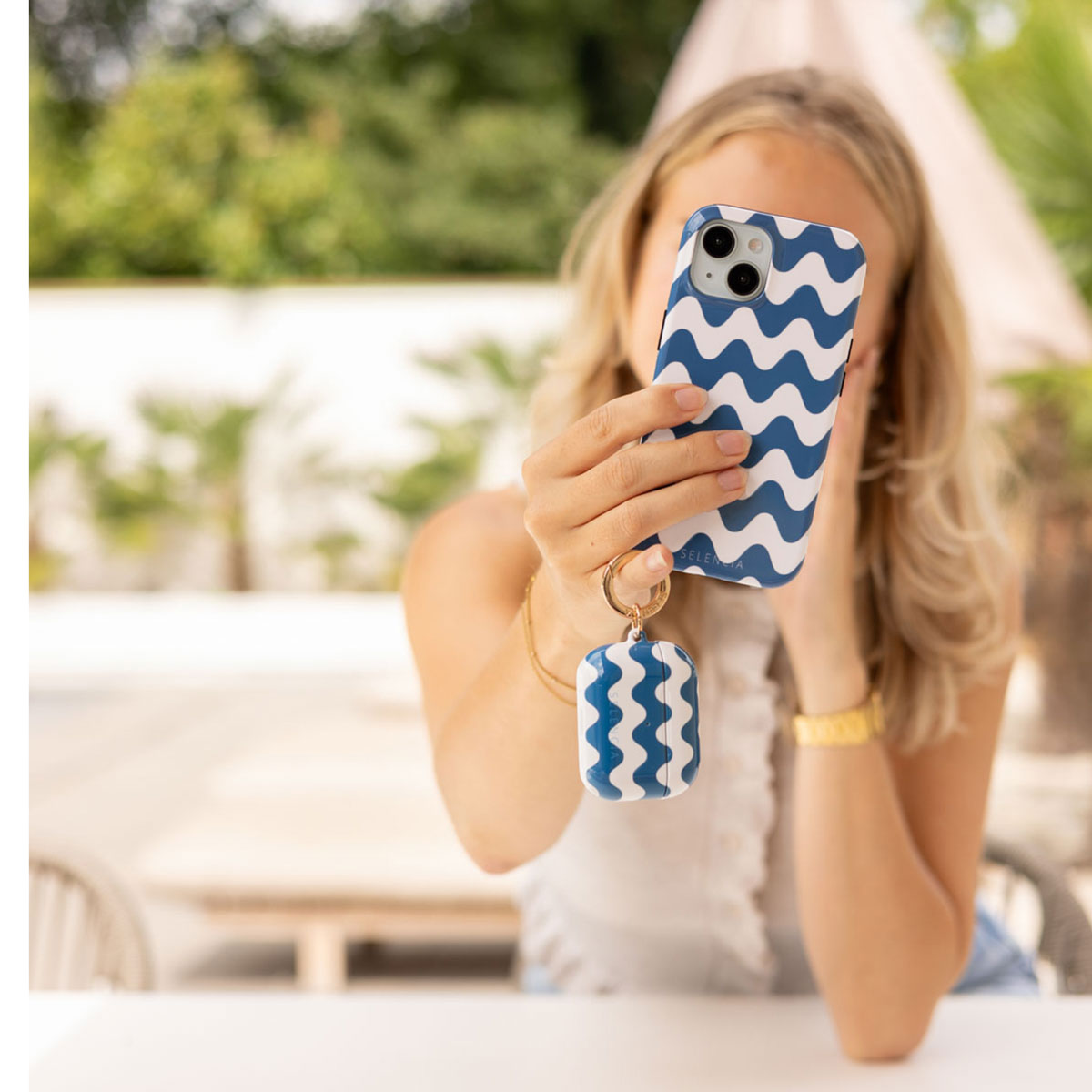 Een vrouw houdt een telefoon vast met een blauw golfpatroon. Een bijpassende AirPods-hoes hangt eronder. De achtergrond toont groen en een parasol.