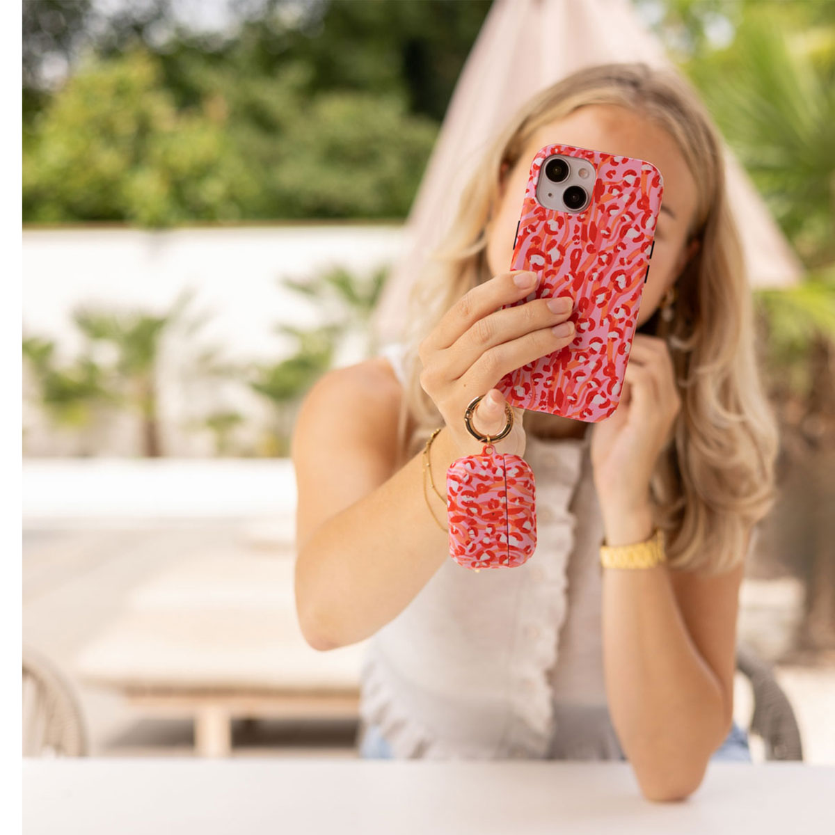Blonde vrouw die een selfie maakt met een roze telefoon met patroon en een AirPods-hoesje. Ze is buiten, draagt een witte top en een gouden horloge.