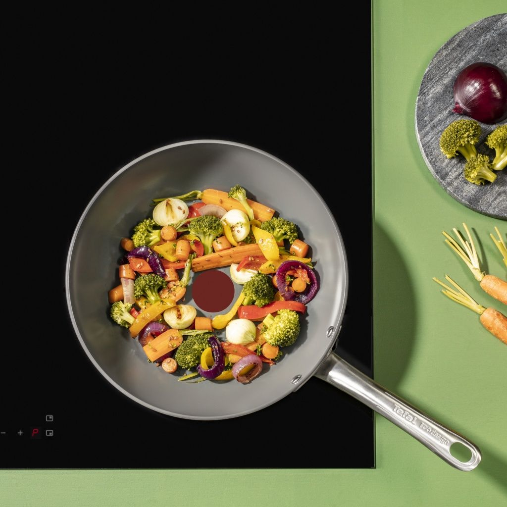 Una padella di verdure su un fornello nero. Broccoli, carote e cipolle sono visibili. Una cipolla rossa e carote sono anche di lato.