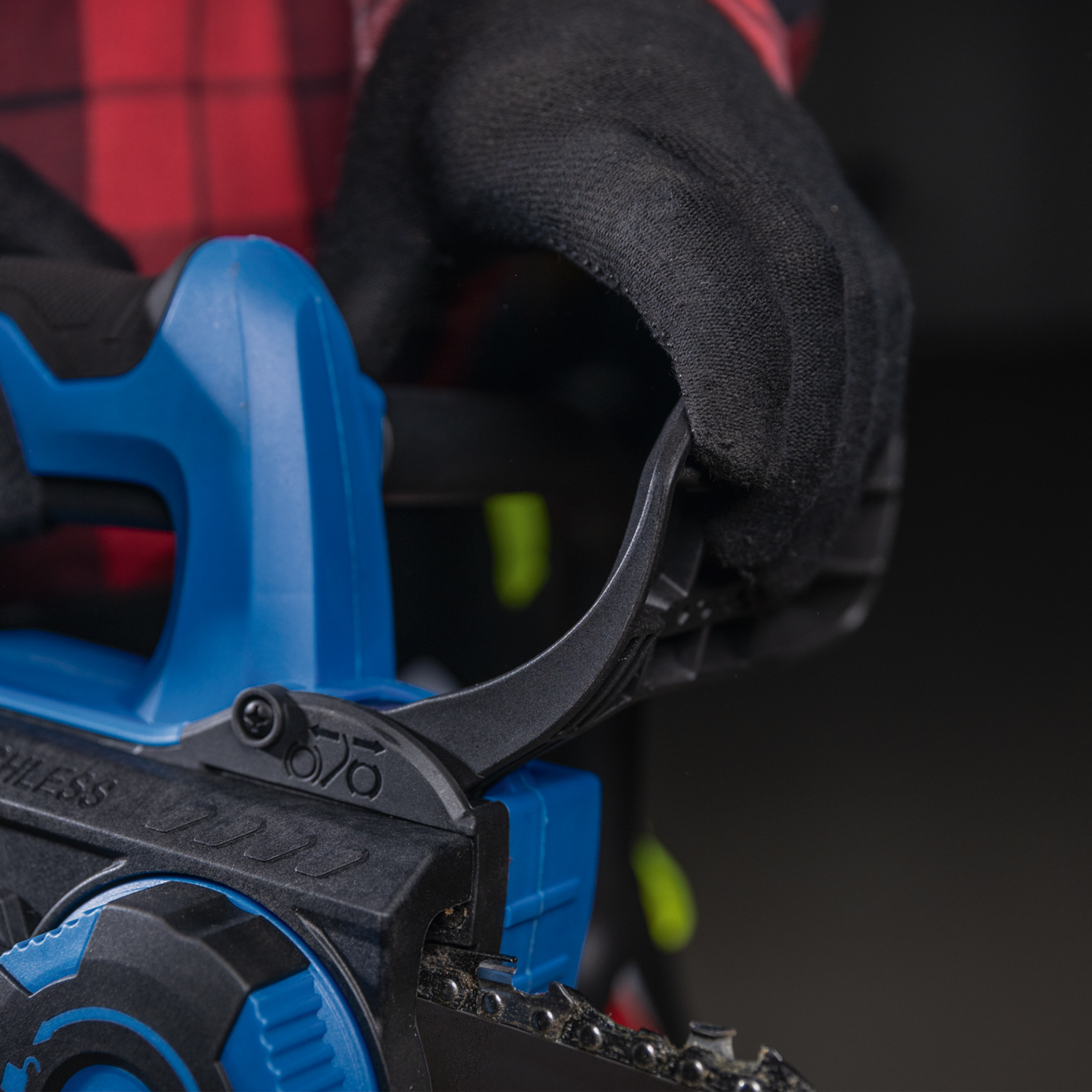 Close-up of a chainsaw. A gloved hand grips the handle. The saw is blue and black. A red and black plaid shirt is visible.