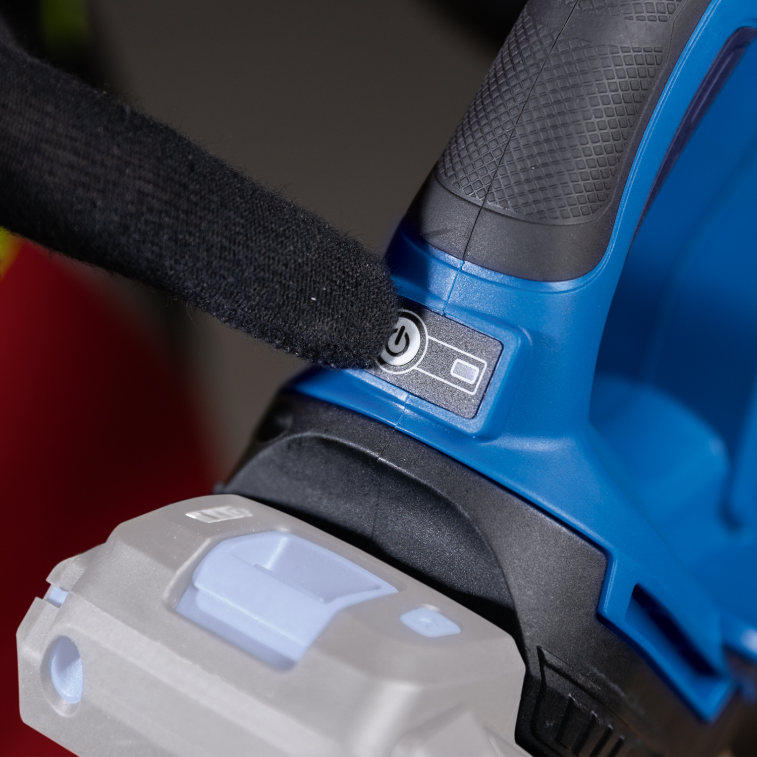 A hand in a black glove presses a power button on a blue and black power tool. A battery is attached below.