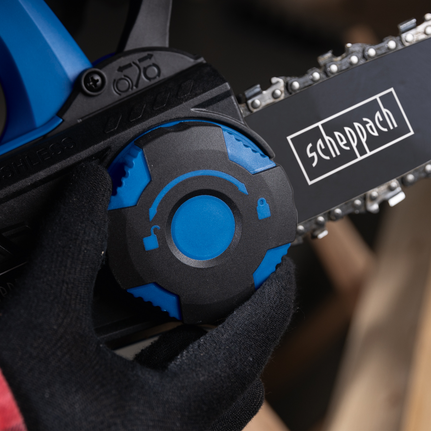 A hand in a black glove holds a blue and black chainsaw. The blade has the 'scheppach' logo. The background is blurry.