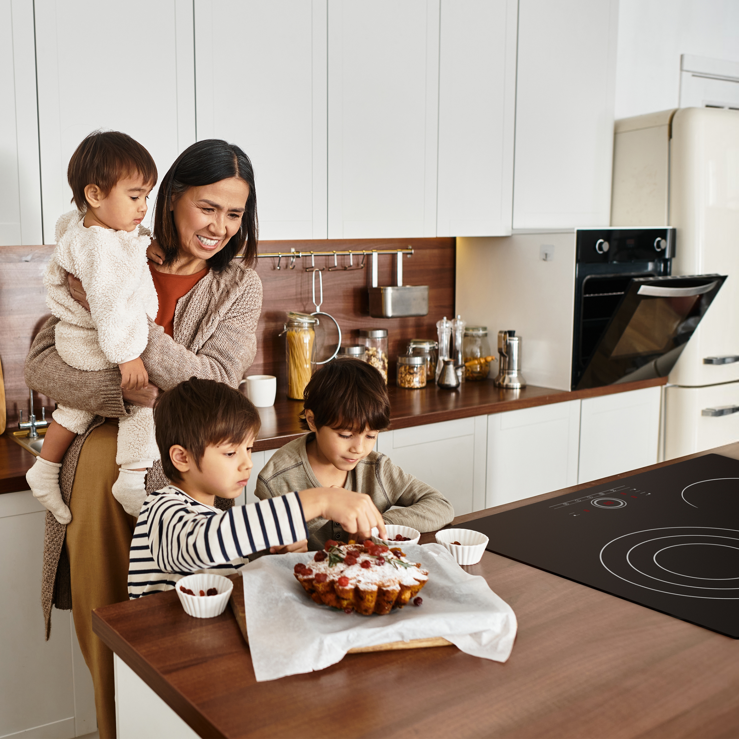 Une femme tient un enfant tandis que deux autres enfants décorent un gâteau dans une cuisine.