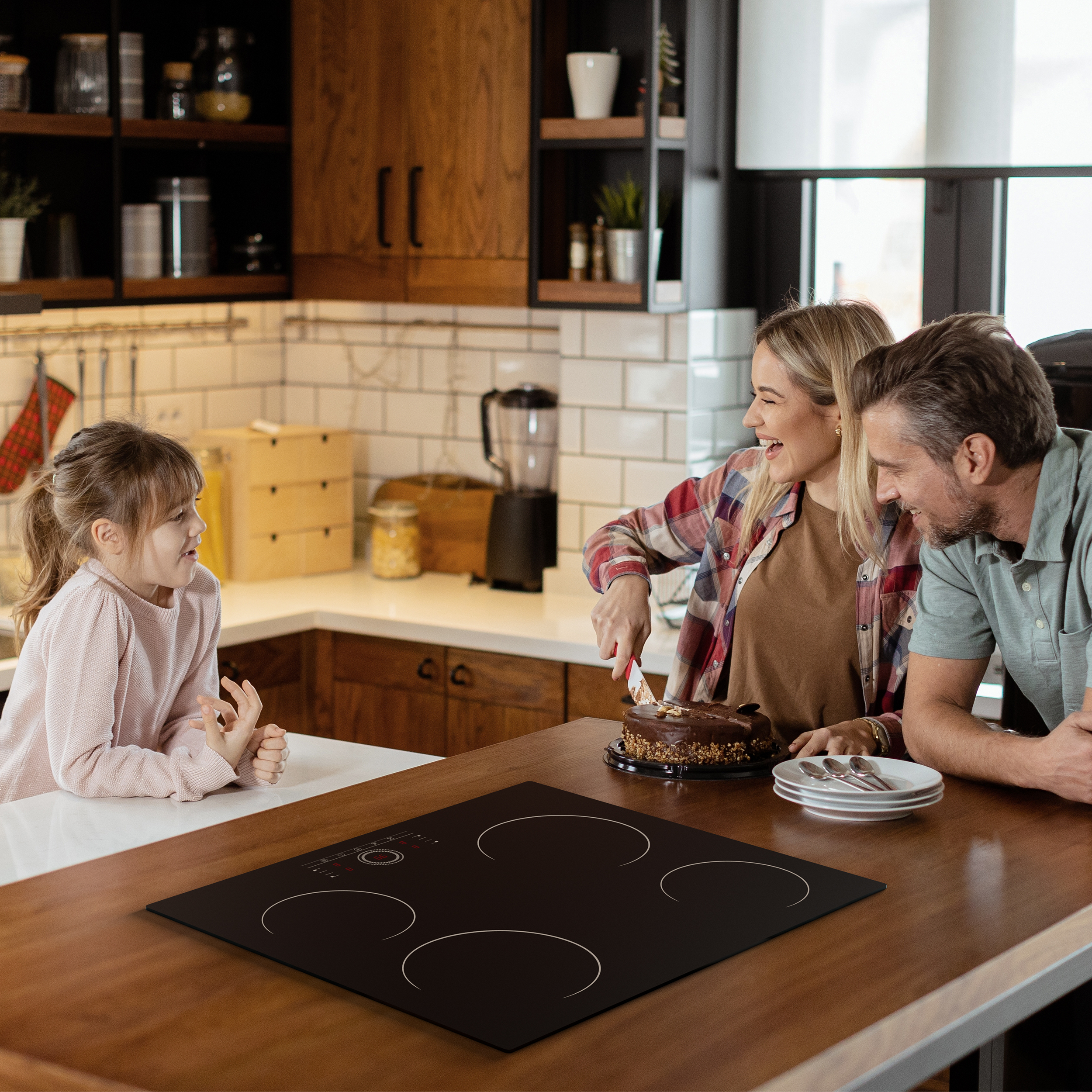 Een gezin viert in een keuken. Een vrouw snijdt een taart. Een kind en een man kijken toe. Borden en een inductiekookplaat zijn zichtbaar.
