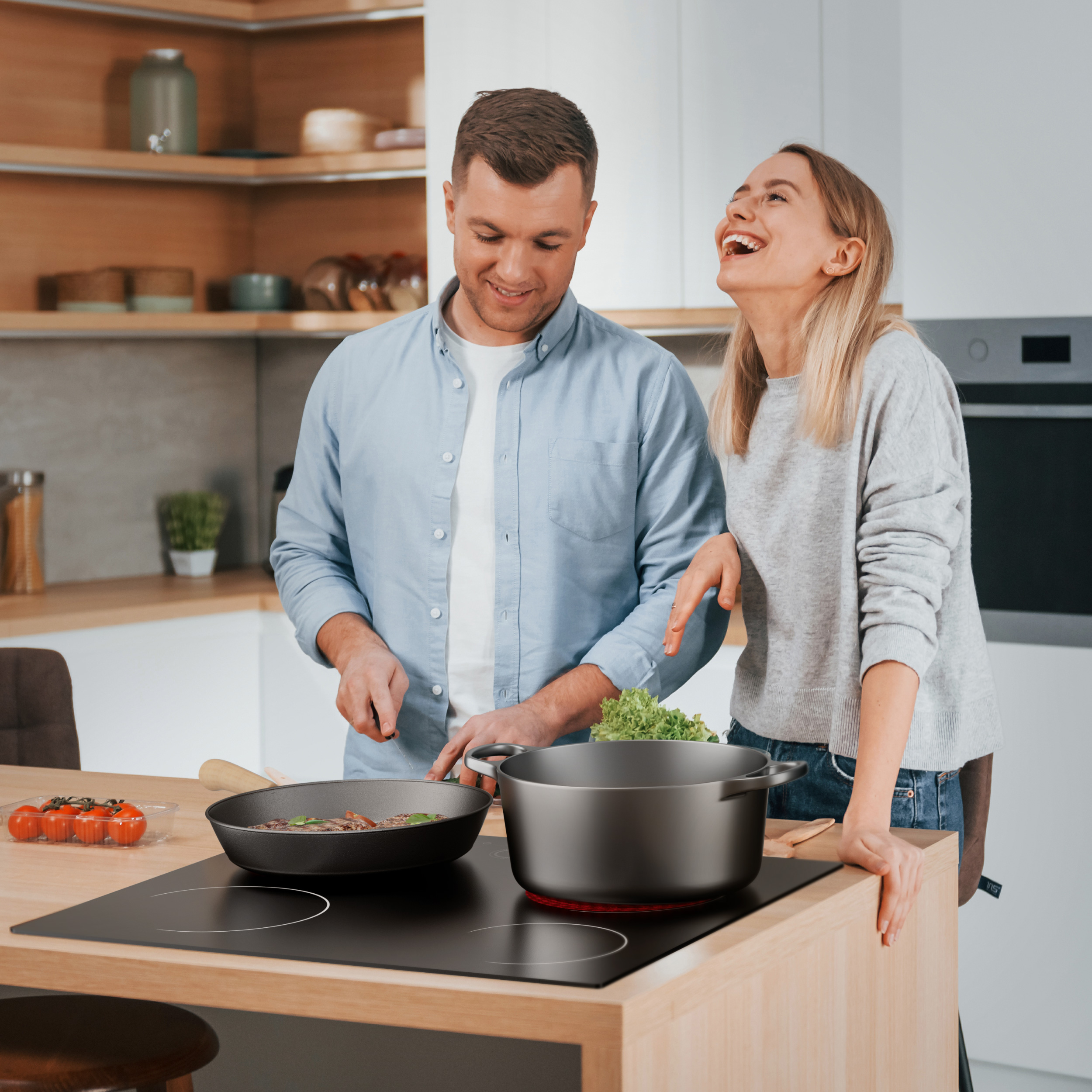 Een stel lacht in een keuken. De man kookt eten in een pan. De vrouw glimlacht, leunend op het aanrecht.