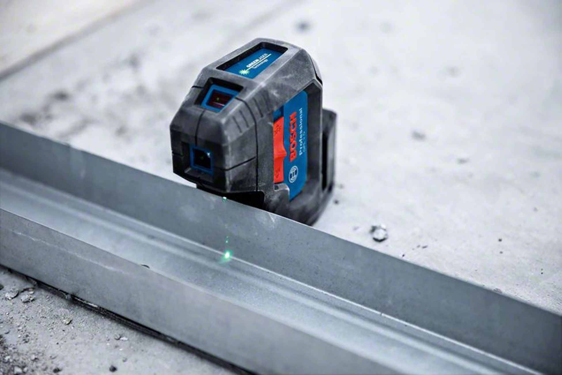 Un nivel láser Bosch proyecta una línea verde sobre una pista de metal. La herramienta es negra, roja y azul, y descansa sobre una superficie de hormigón.