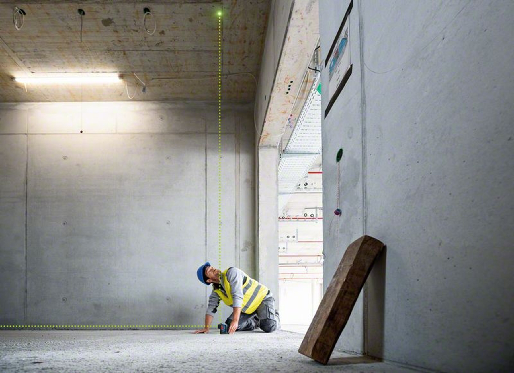Un trabajador de la construcción se arrodilla en el suelo, mirando un nivel láser. Paredes y techo de hormigón. Una viga de madera apoyada en una pared.