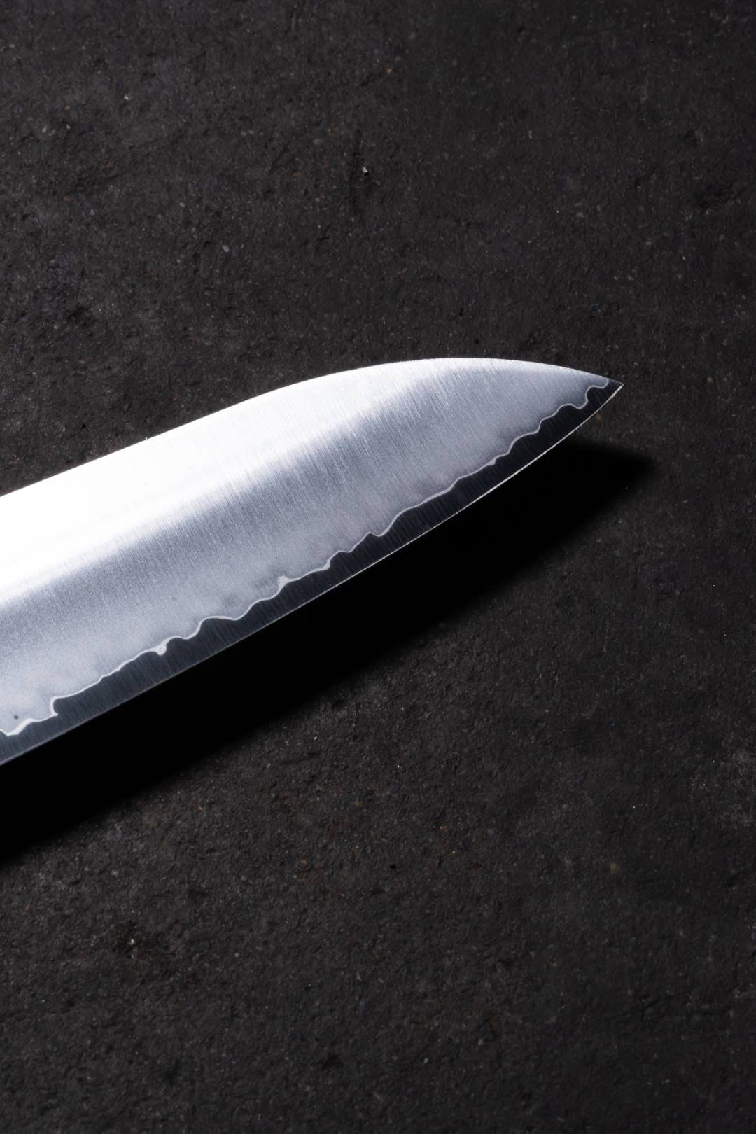 Nahaufnahme einer gezahnten Messerklinge auf dunkler, strukturierter Oberfläche. Die silberne Klinge kontrastiert mit dem schwarzen Hintergrund.