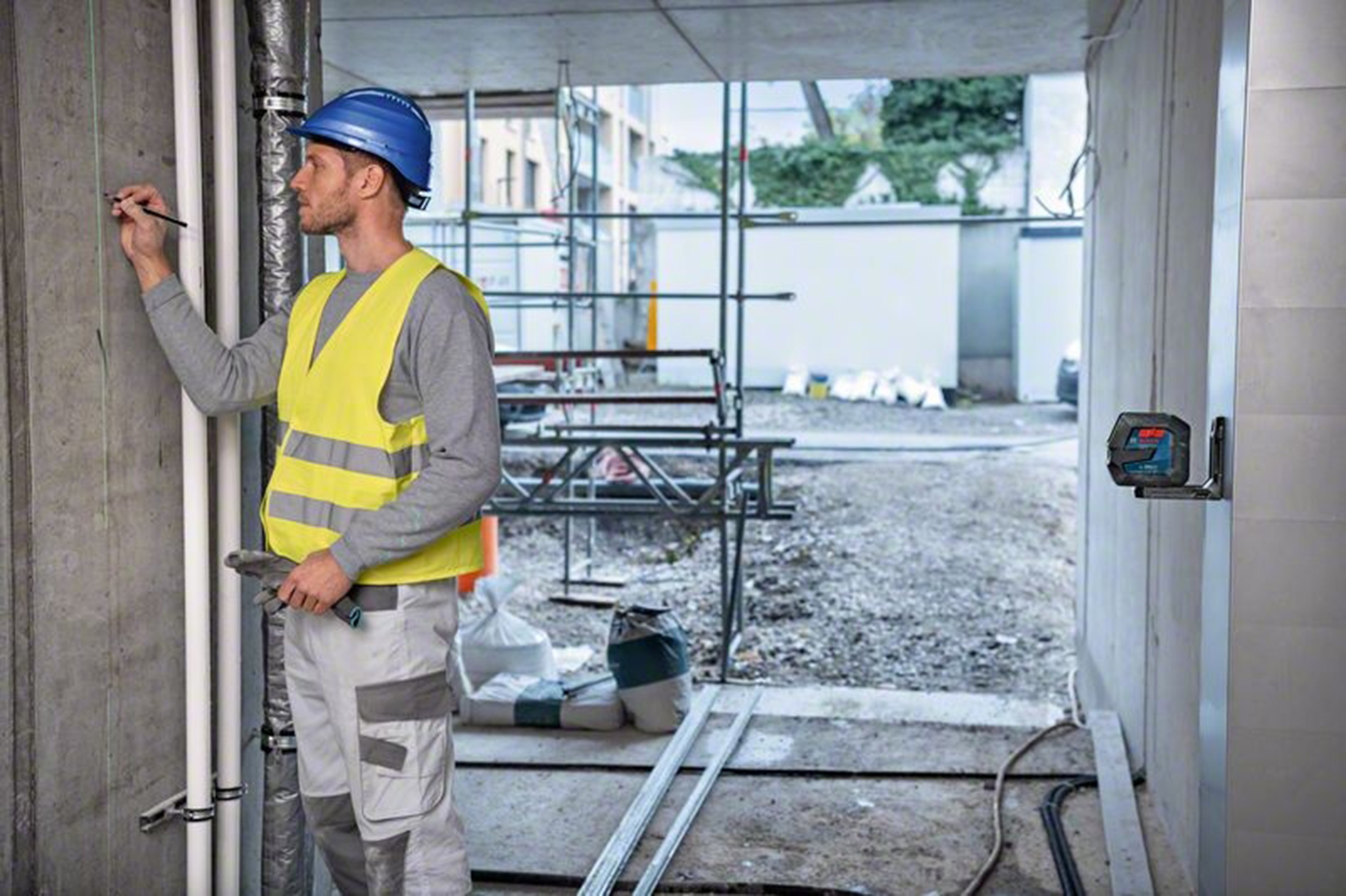 Un operaio edile con casco blu e gilet giallo segna un muro. Livella laser montata. Sfondo cantiere.