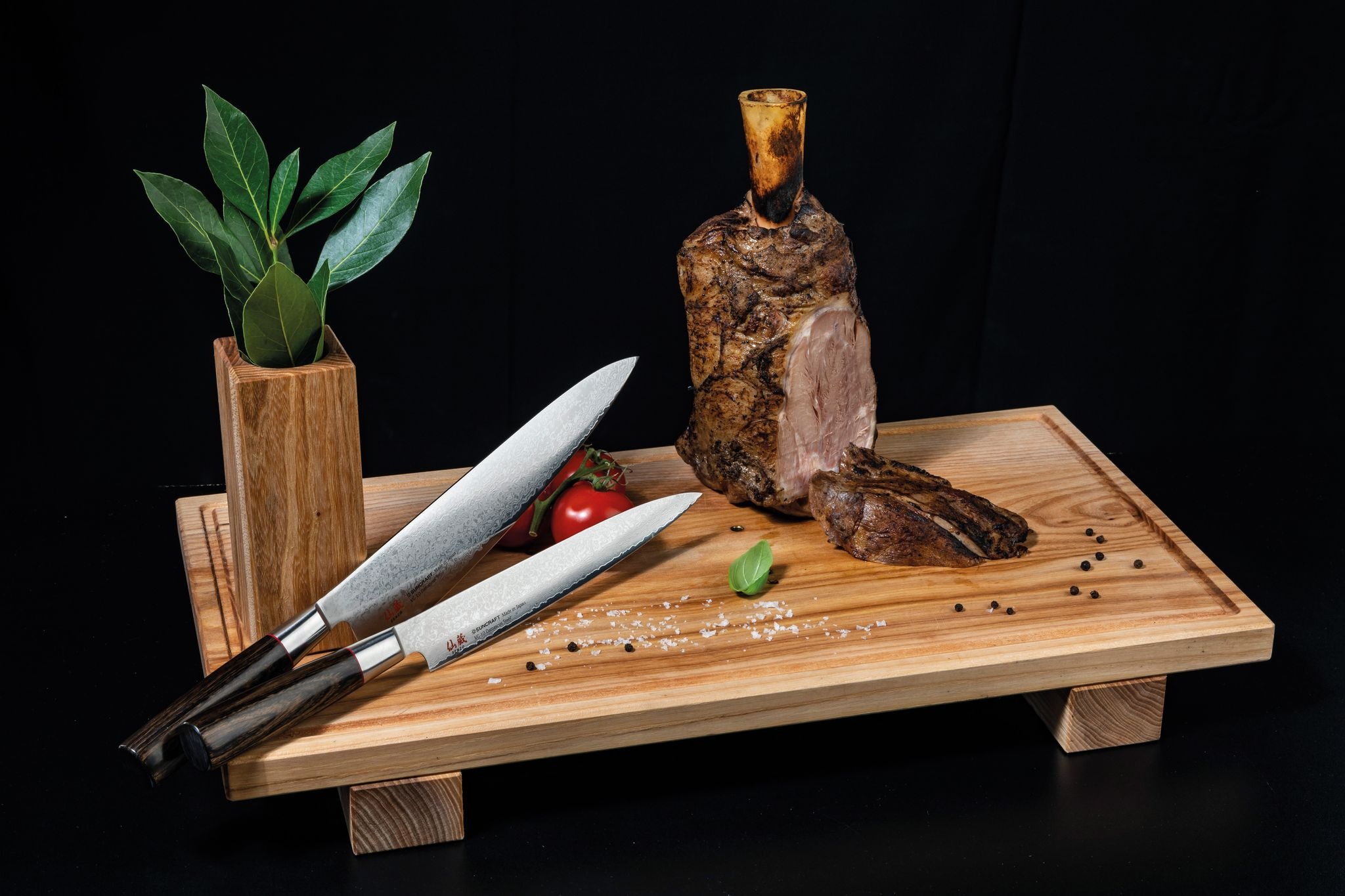 Ein Holzbrett mit gebratenem Fleisch, Messern, Tomaten und Kräutern. Schwarzer Hintergrund.