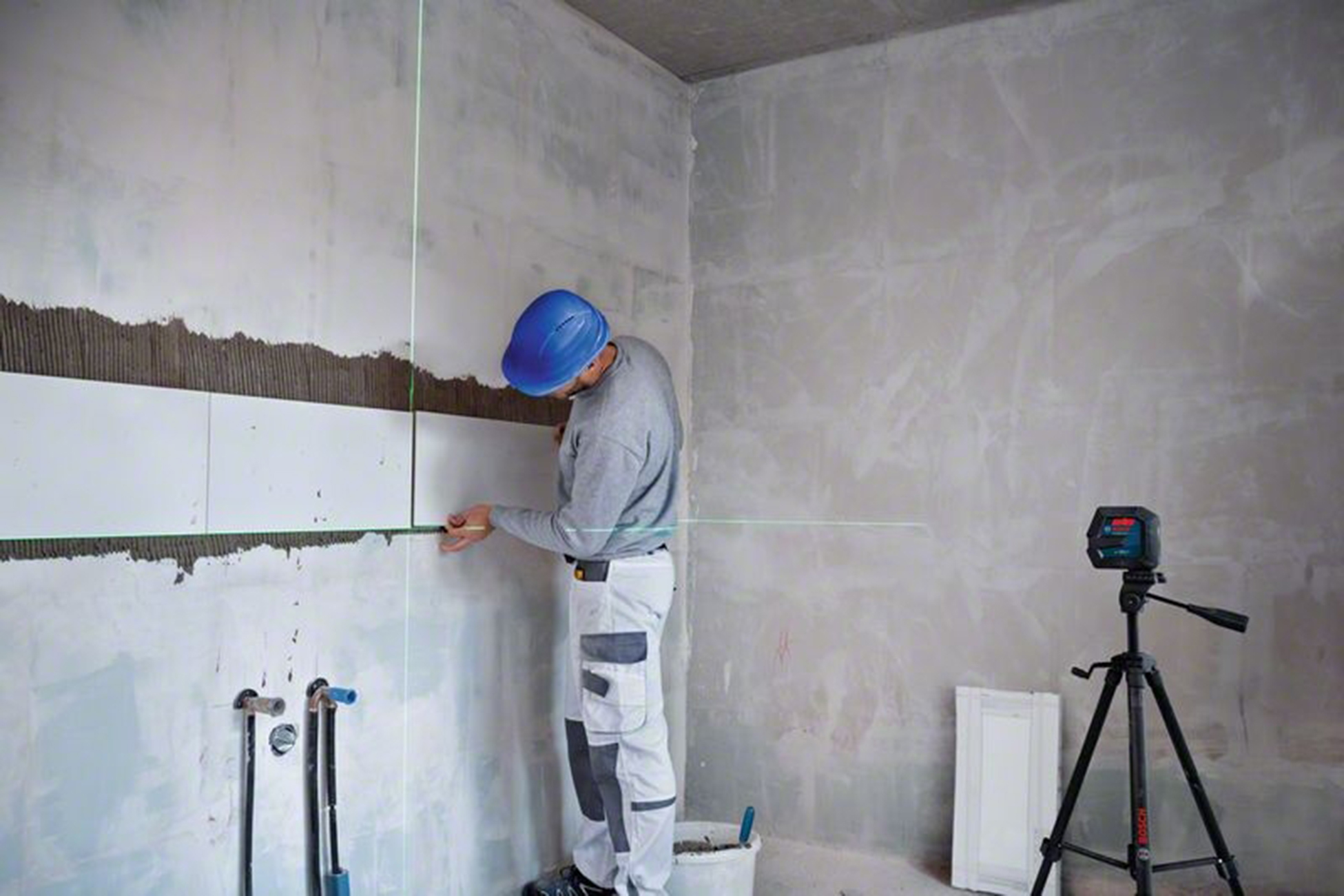 Un uomo con un casco blu sta piastrellando un muro. Si usa un livello laser. Il muro è incompiuto e alcune piastrelle sono già posizionate.