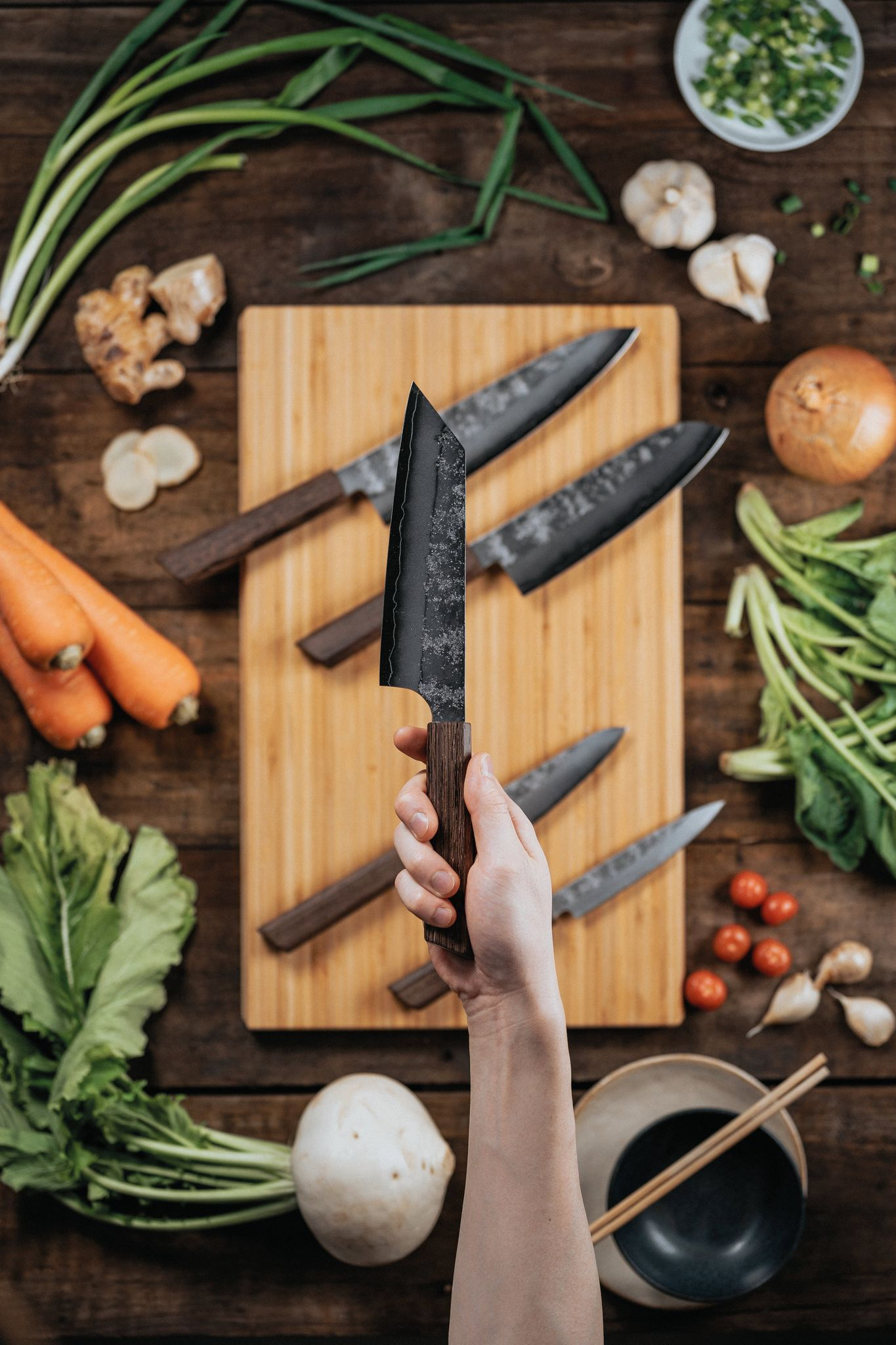 Hand hält ein Messer über einem Holzschneidebrett, umgeben von Gemüse, darunter Karotten, Zwiebeln und Grünzeug. Eine Schüssel und Essstäbchen sind vorhanden.