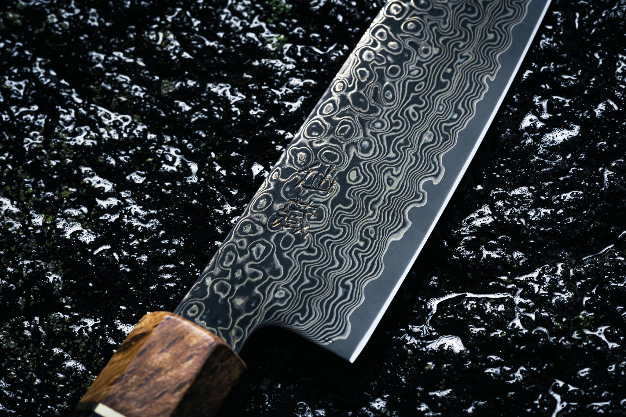 Nahaufnahme einer gemusterten Messerklinge mit Holzgriff. Die Klinge hat ein wellenförmiges Design. Der Hintergrund ist dunkel und strukturiert.