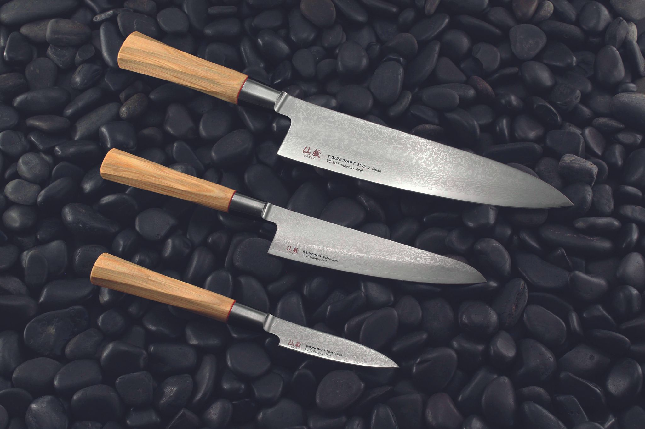 Drei japanische Küchenmesser mit Holzgriffen liegen auf schwarzen Kieselsteinen. Die Klingen sind silbern und zeigen ein Damastmuster.