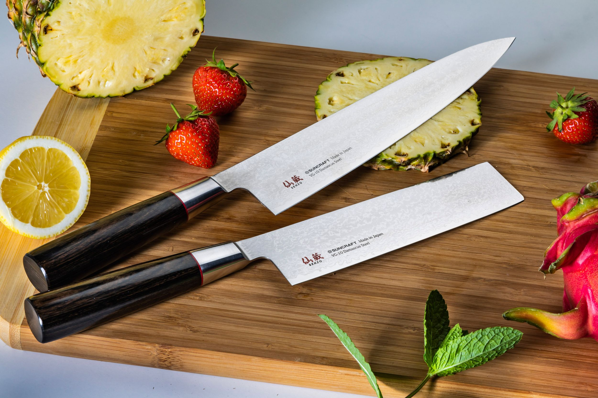 Zwei Messer auf einem Holzbrett mit Früchten: Ananas, Erdbeeren, Zitrone, Drachenfrucht und Minze.