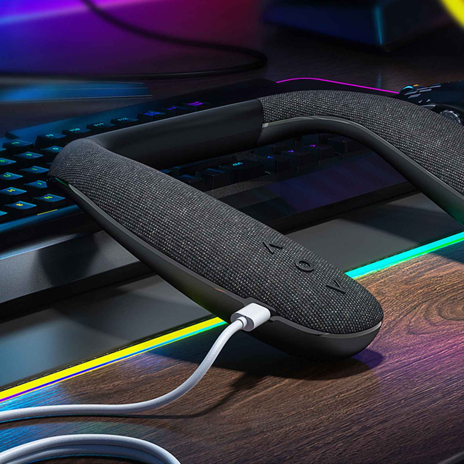 Ein schwarzer Nackenbügellautsprecher mit weißem Ladekabel auf einem Holzschreibtisch in der Nähe einer Tastatur.