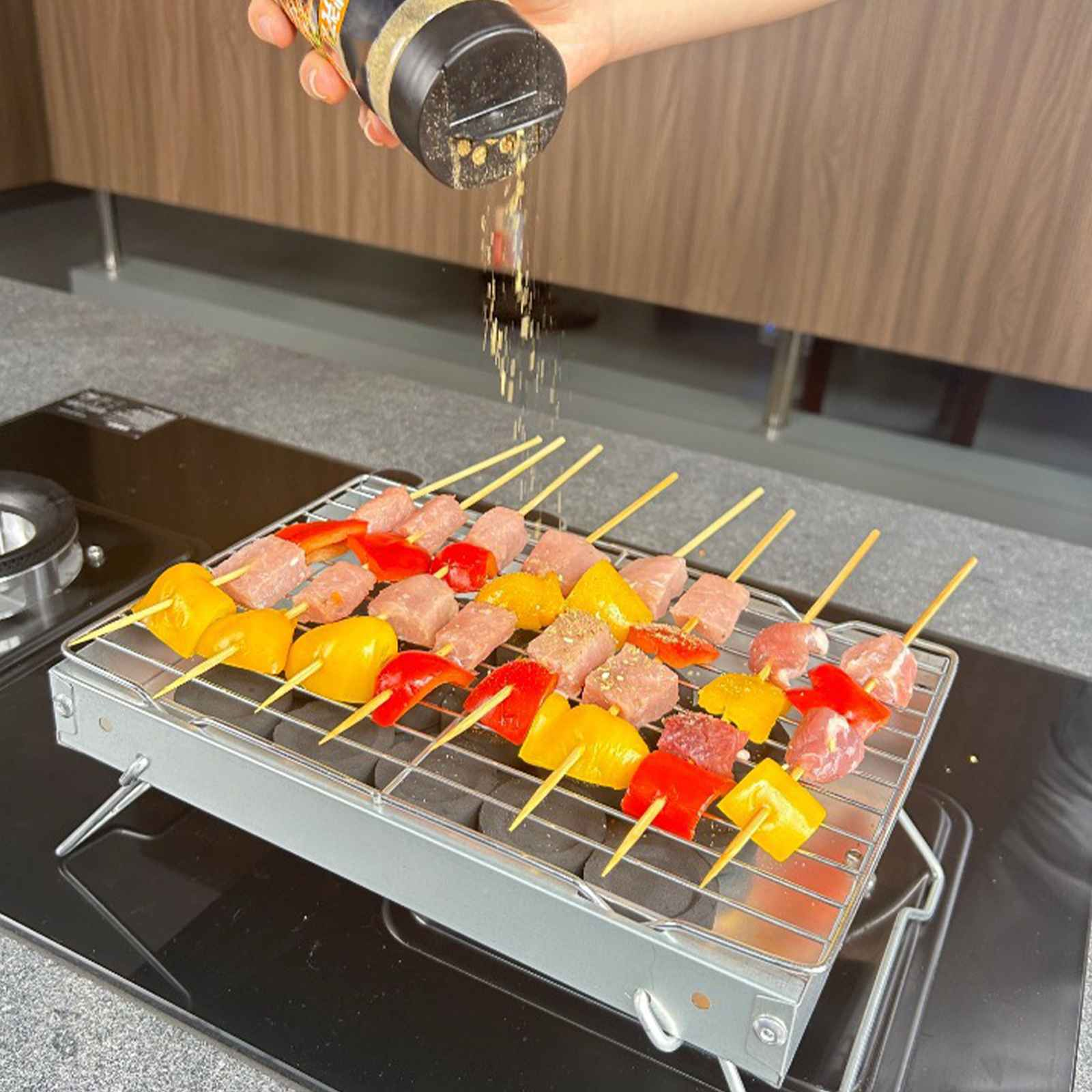 Hand würzt Spieße aus Fleisch und Gemüse auf einem kleinen Grill. Gelbe, rote Paprika und Fleisch sind sichtbar.