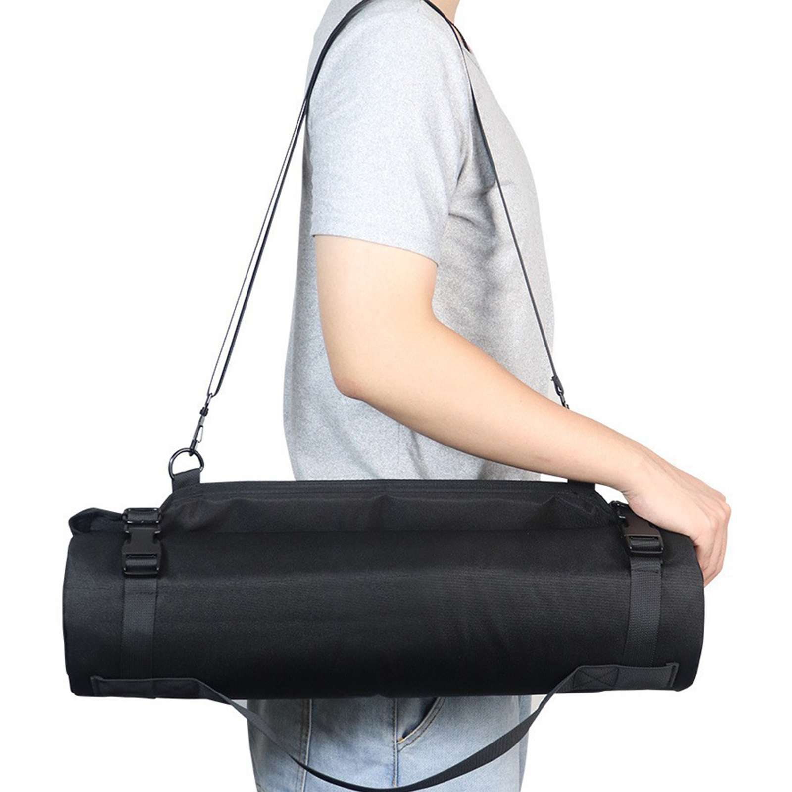 Eine Person trägt eine schwarze zylindrische Tasche über der Schulter. Tasche hat schwarze Riemen und Schnallen. Person trägt graues Hemd und Jeans.