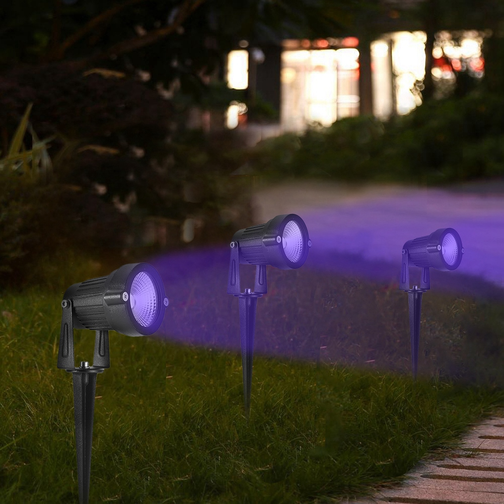 Drei Outdoor-Scheinwerfer mit lila Licht auf dem Rasen. Im Hintergrund ist ein Haus zu sehen, und die Szene ist beleuchtet.