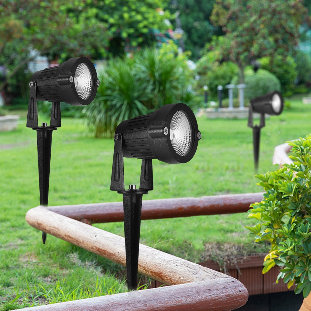 Drei schwarze Scheinwerfer in einem Garten. Sie stehen auf dem Rasen, in der Nähe eines Holzzauns, mit Bäumen im Hintergrund.