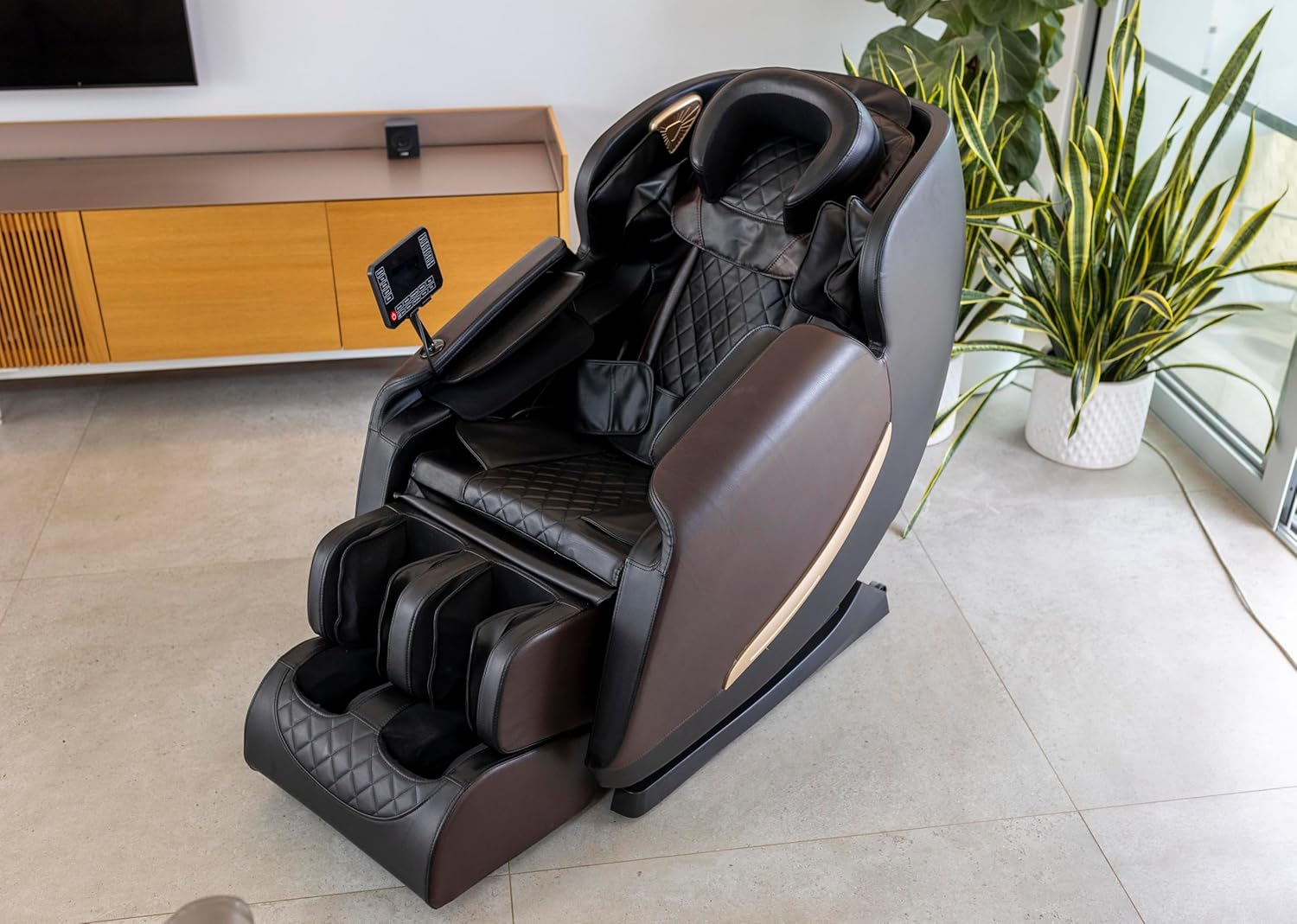 Un sillón de masaje moderno en una habitación. Es negro y marrón con un panel de control. Hay plantas y muebles en el fondo.