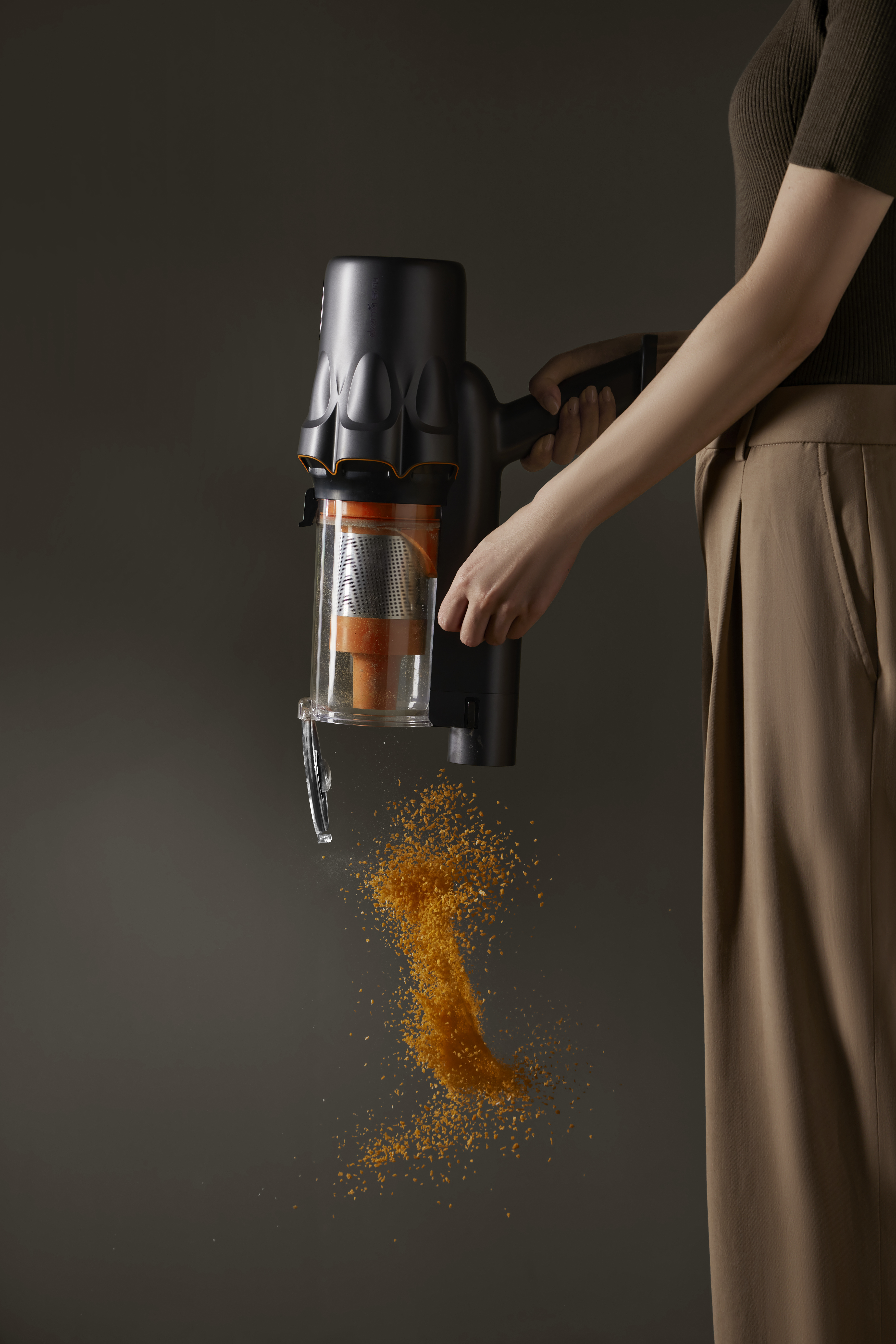 Osoba trzyma czarny odkurzacz, opróżniając pomarańczowe śmieci. Tło jest gładkie szare.