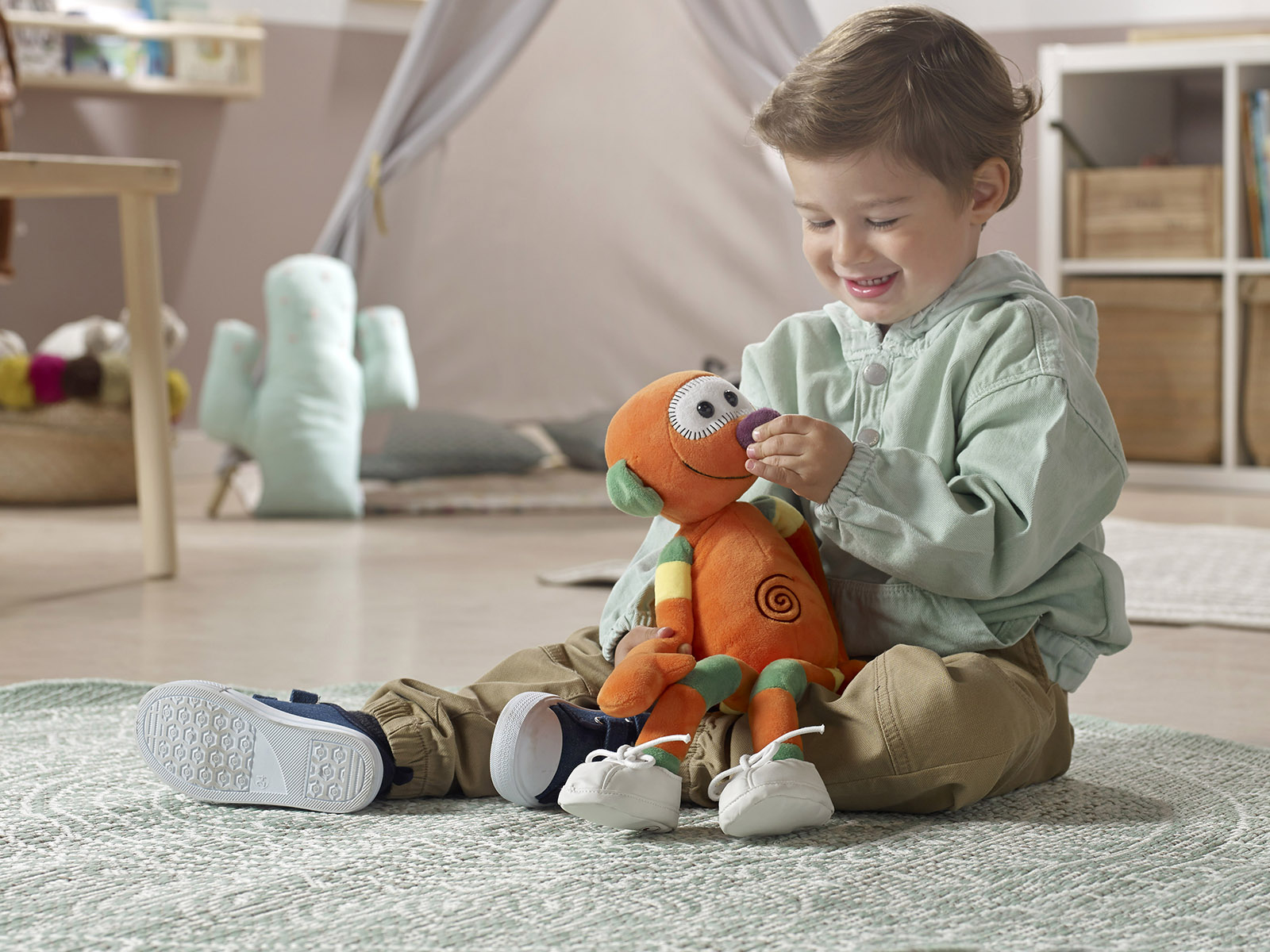 Mały chłopiec uśmiecha się, trzymając pomarańczową pluszową zabawkę. Siedzi na dywanie, w tle namiot i poduszka w kształcie kaktusa.