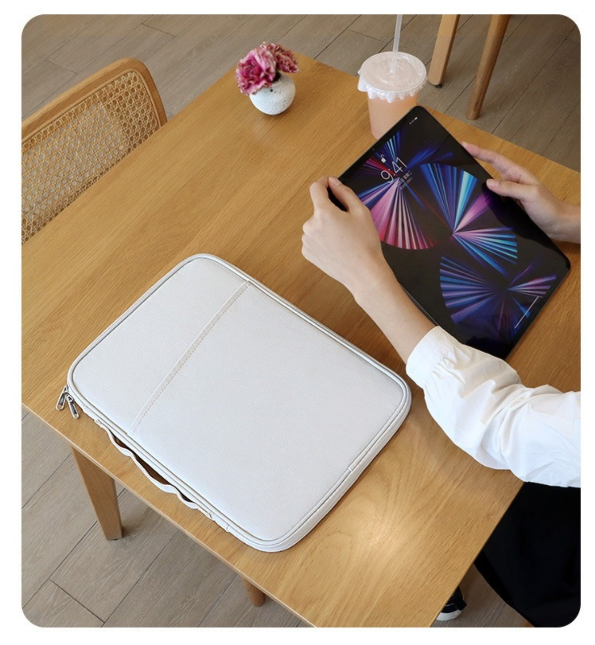 Eine Person hält ein Tablet auf einem Holztisch. Es gibt eine weiße Hülle, ein Getränk und Blumen. Links ist ein Stuhl.