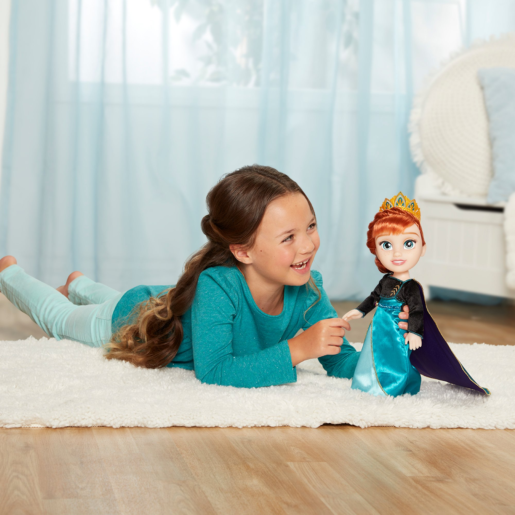 Una niña está tumbada en una alfombra, sosteniendo una muñeca de Anna. La muñeca tiene una corona y un vestido azul. La niña sonríe.