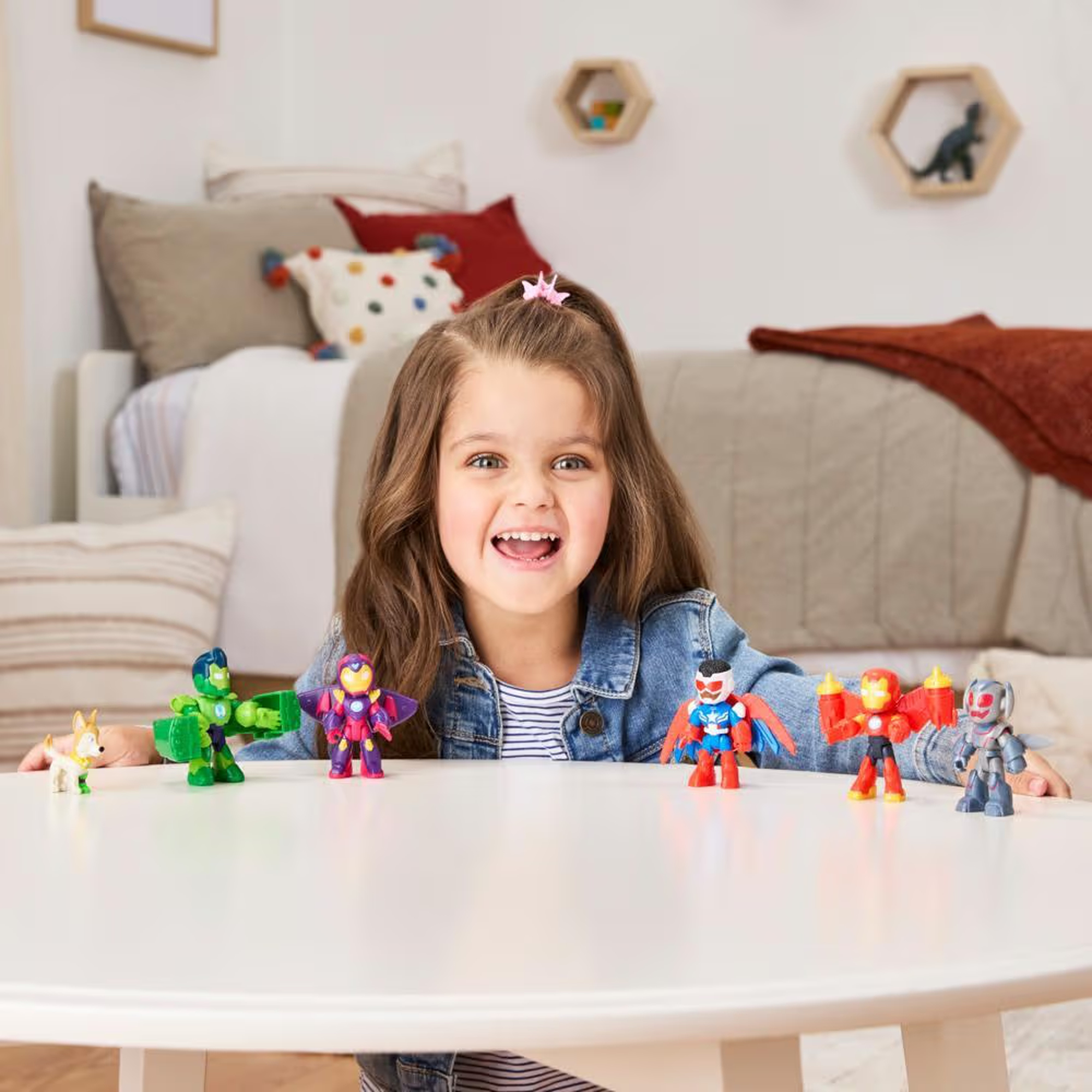 Una niña sonriente con chaqueta vaquera y juguetes en una mesa blanca. El fondo incluye una cama y estantes decorativos.