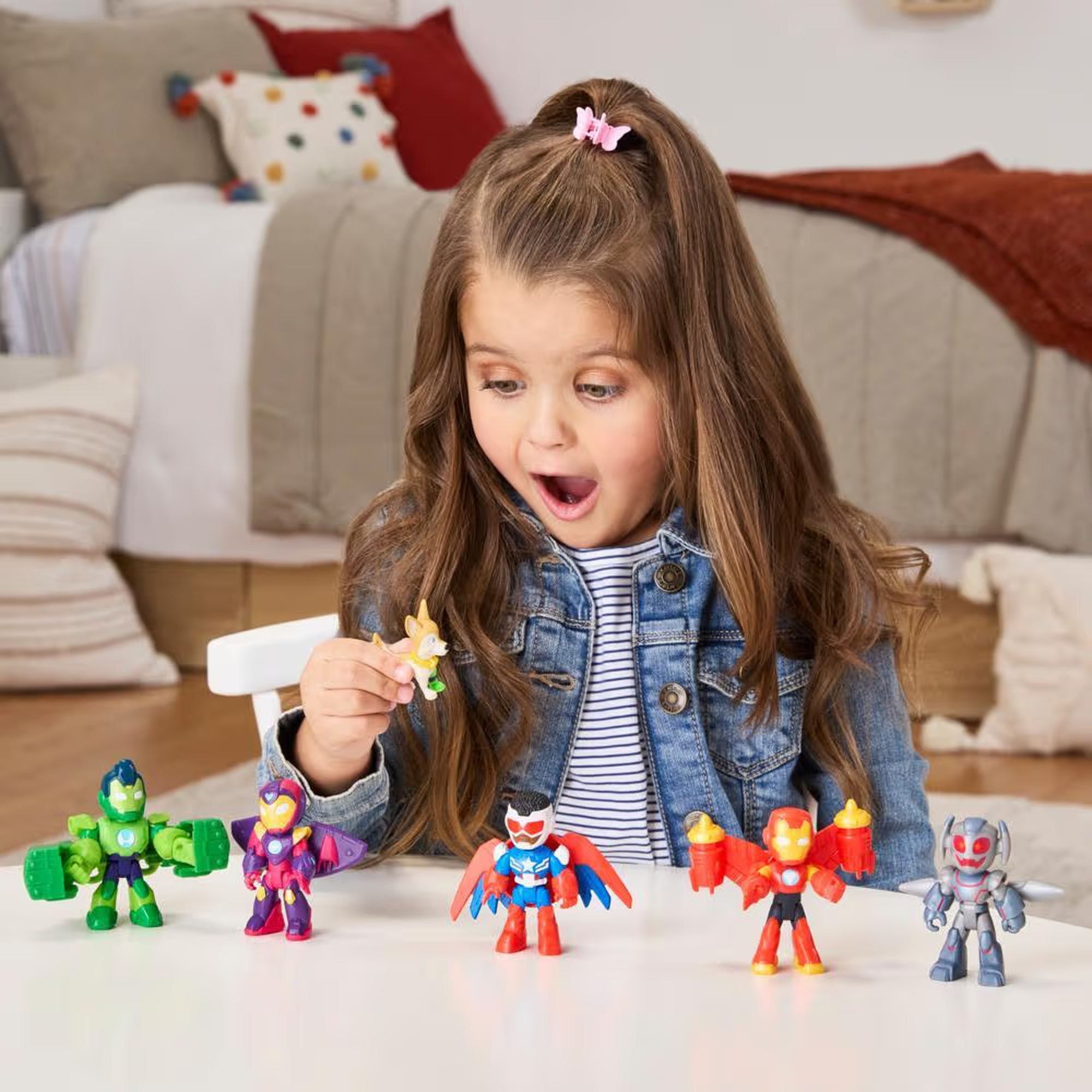 Niña con la boca abierta mirando un juguete. Juguetes de superhéroes sobre una mesa blanca. El fondo es una cama.