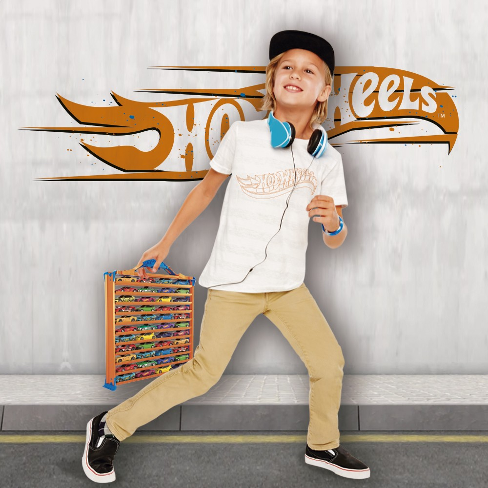 Ein Junge mit weißem Hemd, schwarzer Mütze und Kopfhörern rennt, während er eine Hot Wheels-Tasche hält. Der Hintergrund zeigt ein Logo.