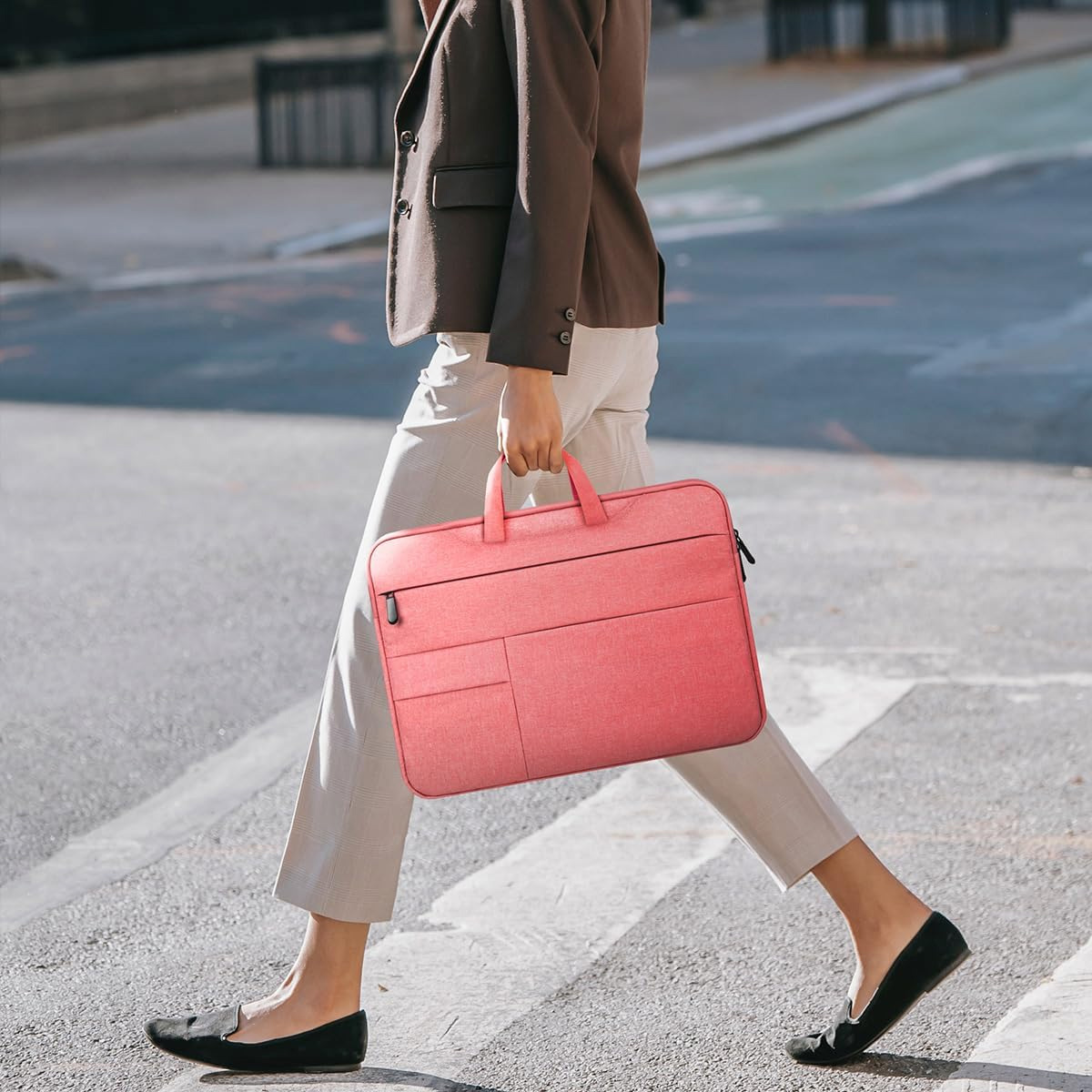 Eine Person überquert einen Zebrastreifen und hält eine pinke Laptoptasche. Sie trägt einen braunen Blazer, graue Hose und schwarze Schuhe. Stadt im Hintergrund.