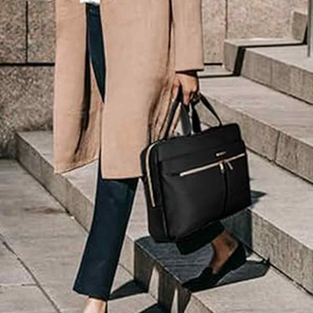 Person geht Treppe runter, hält schwarze Aktentasche. Sie trägt einen beigefarbenen Mantel, eine marineblaue Hose und schwarze Schuhe.