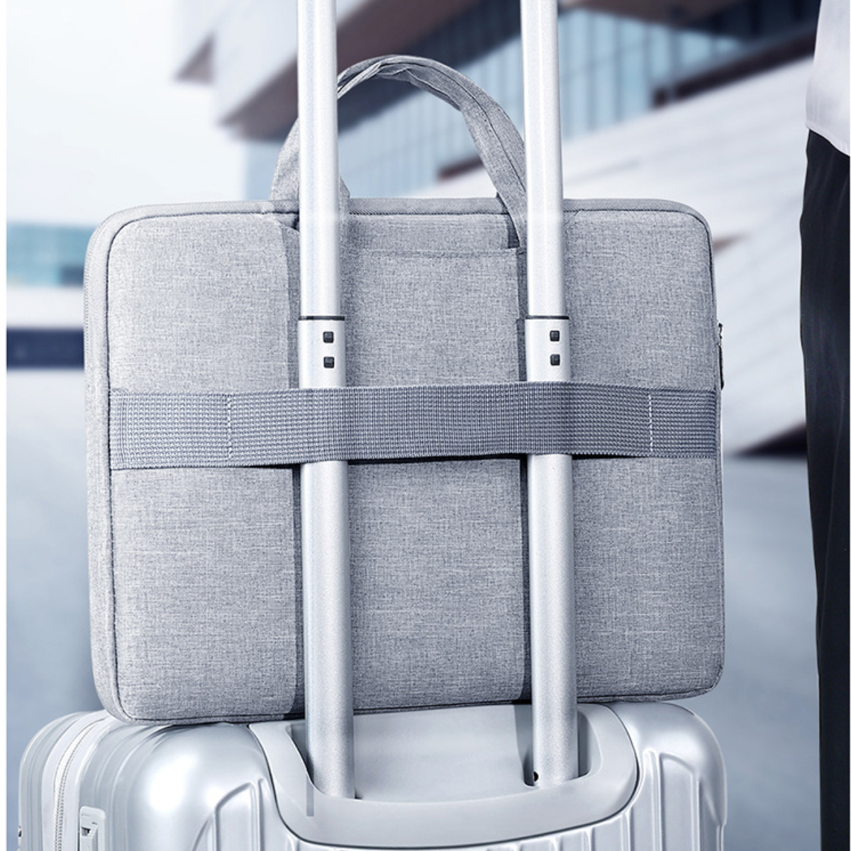 Eine graue Laptoptasche ist mit einem Gurt an einem silbernen Koffer befestigt. Die Tasche hat einen Griff. Eine Person steht in der Nähe.