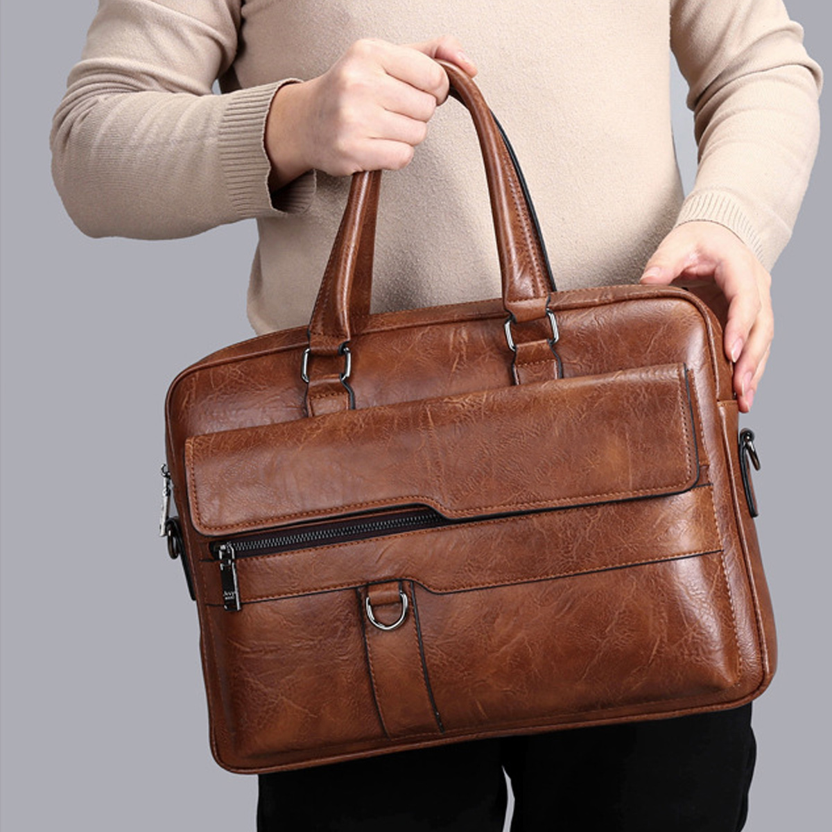 Eine Person hält eine braune Lederaktentasche. Die Tasche hat einen Griff, eine Vordertasche mit Reißverschluss und einen grauen Hintergrund.