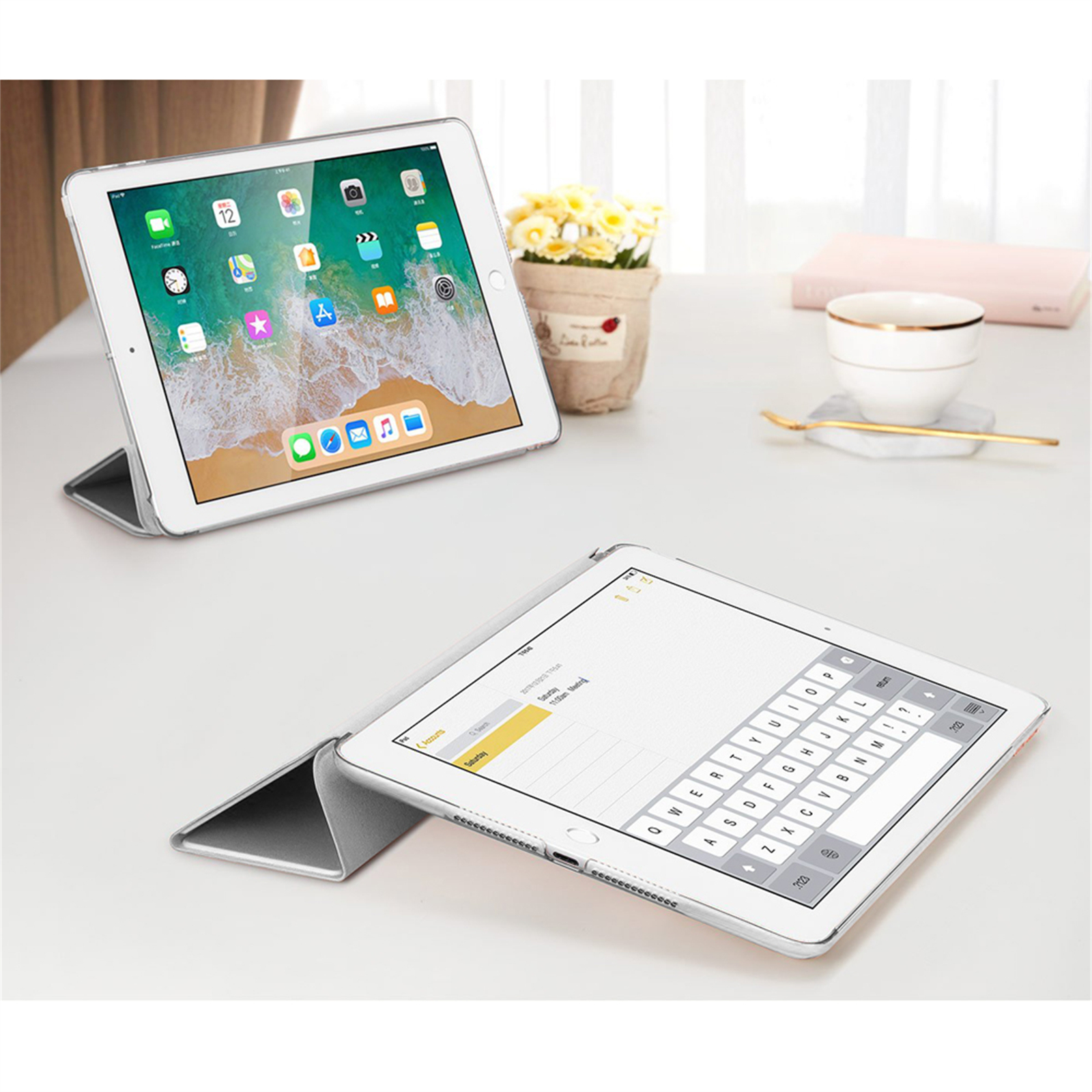 Zwei iPads auf einem weißen Tisch. Eines zeigt Apps, das andere eine Tastatur. Blumentopf, Tasse und Buch sind ebenfalls vorhanden.