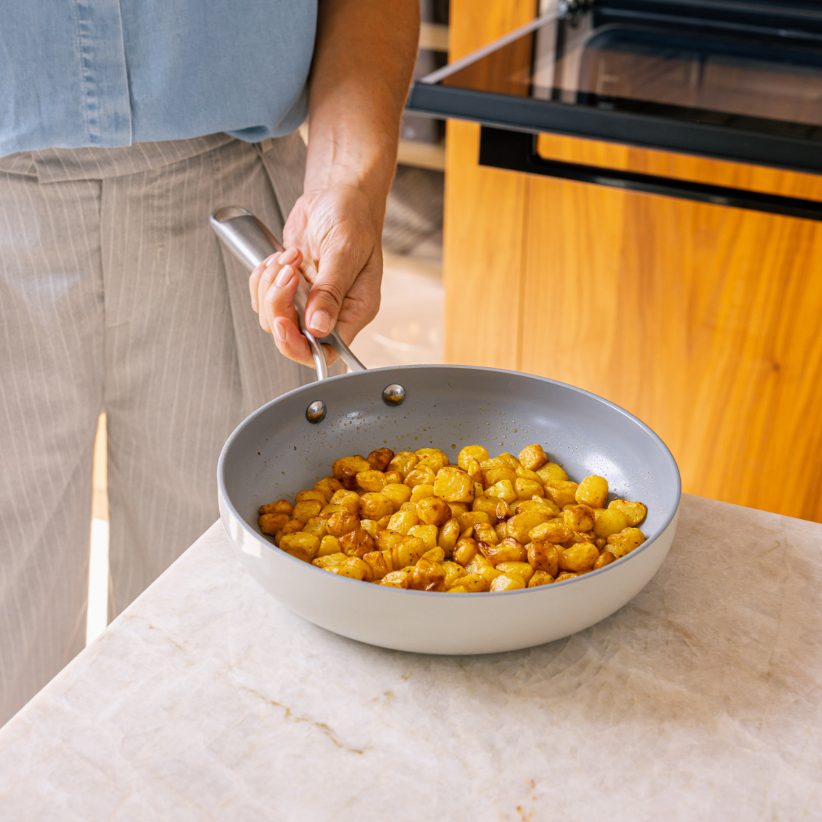 A person holds a pan of golden-brown potatoes. The pan rests on a marble countertop. A stove and wooden cabinet are in the background.