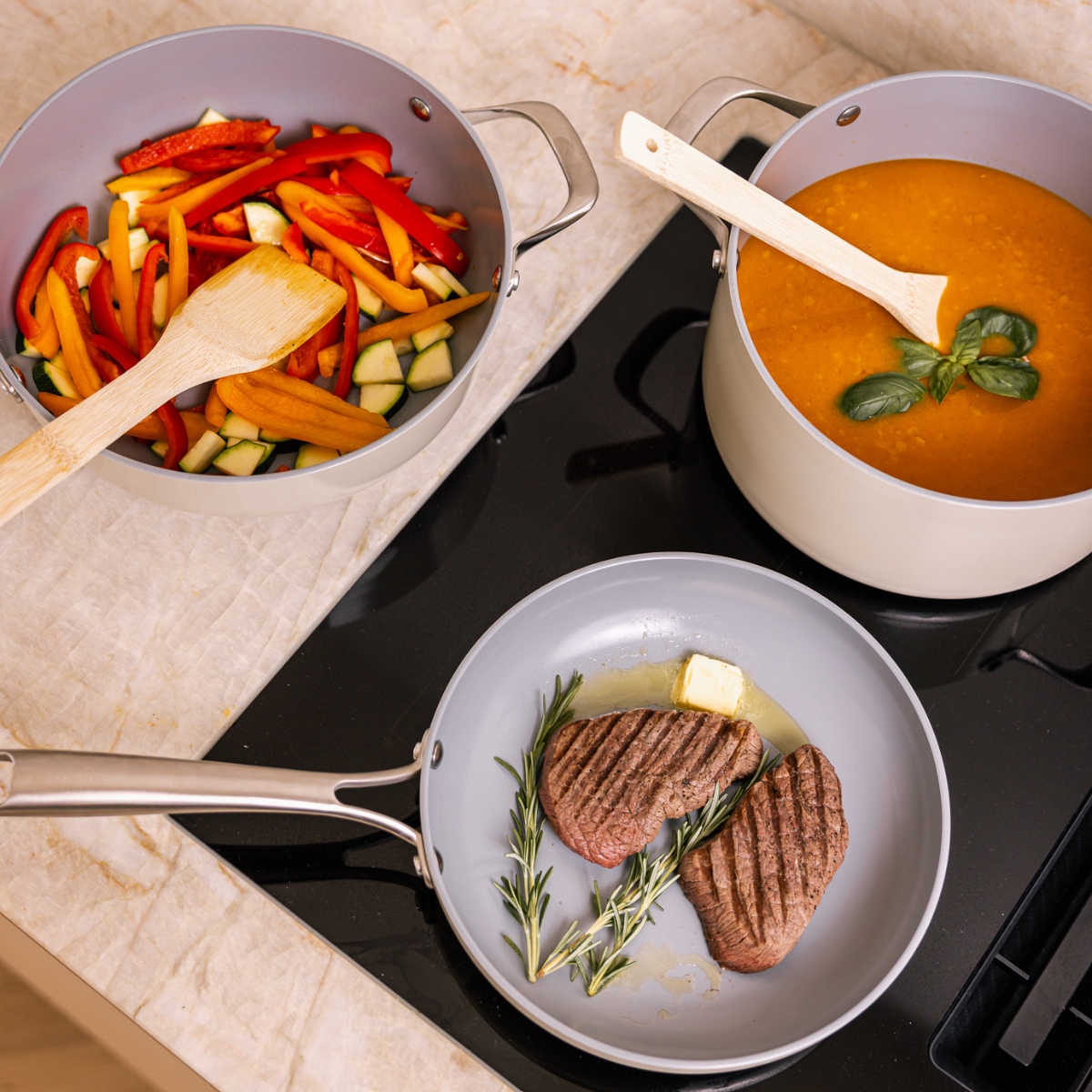 Pans with food on a stove. Steak, soup, and vegetables are visible. Wooden spoons rest in the pans. Butter and herbs surround the steak.