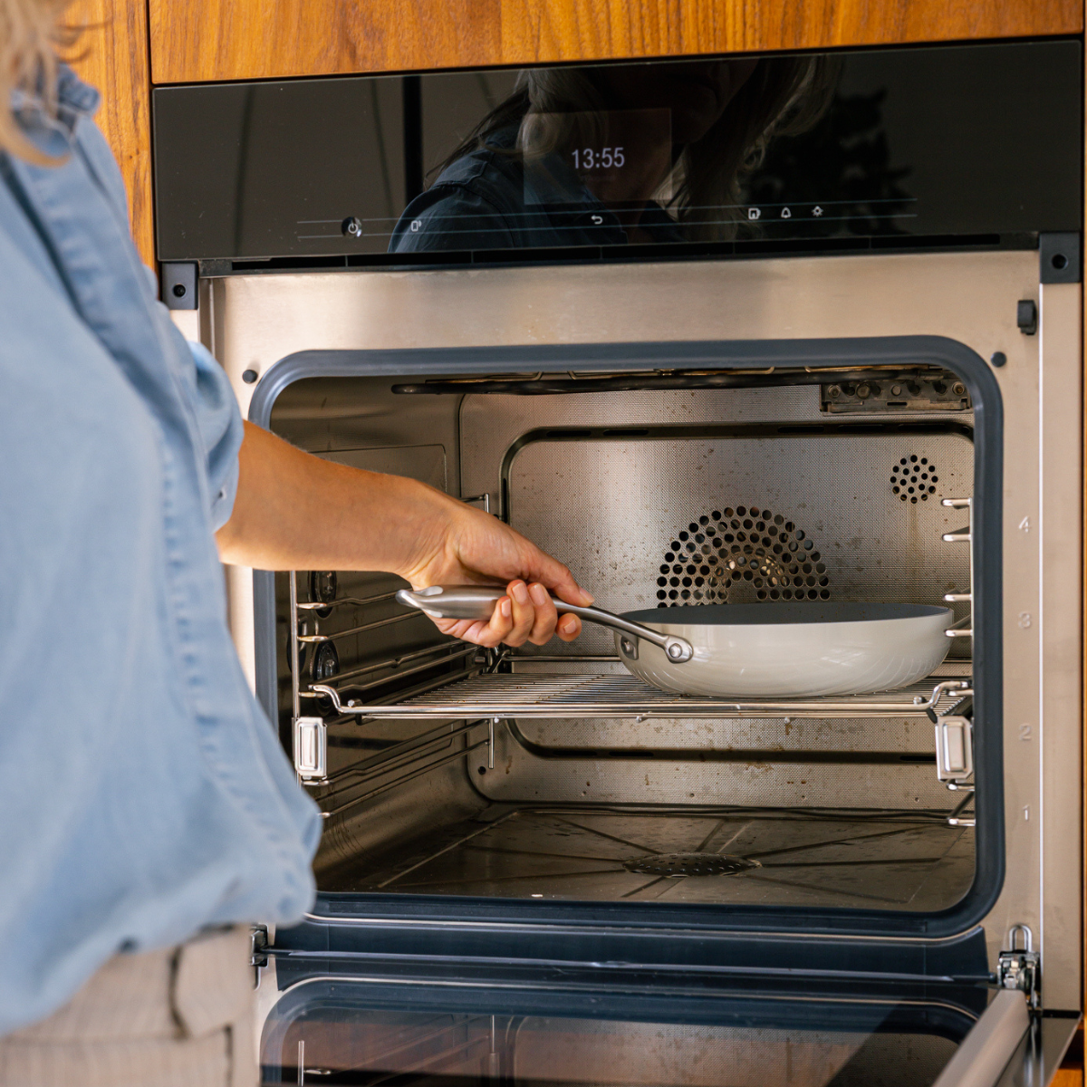 A person places a white pan into a stainless steel oven. The oven door is open. The time 13:55 is displayed on the oven.