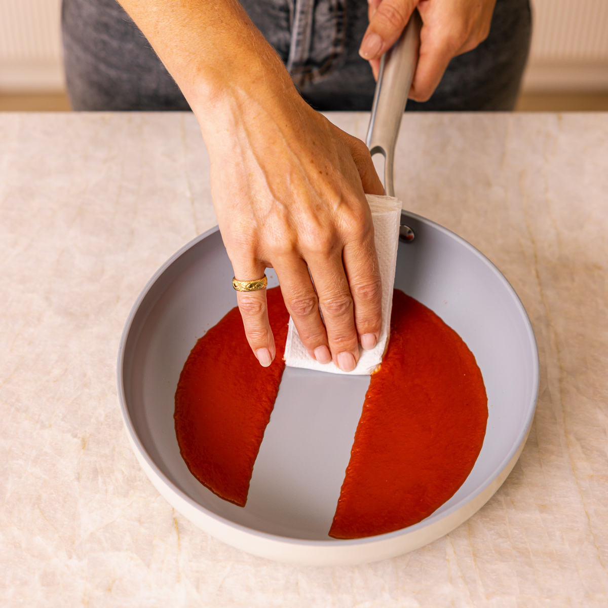 A person wipes a red substance in a pan with a paper towel. The pan is gray with a white handle. The person wears a ring.