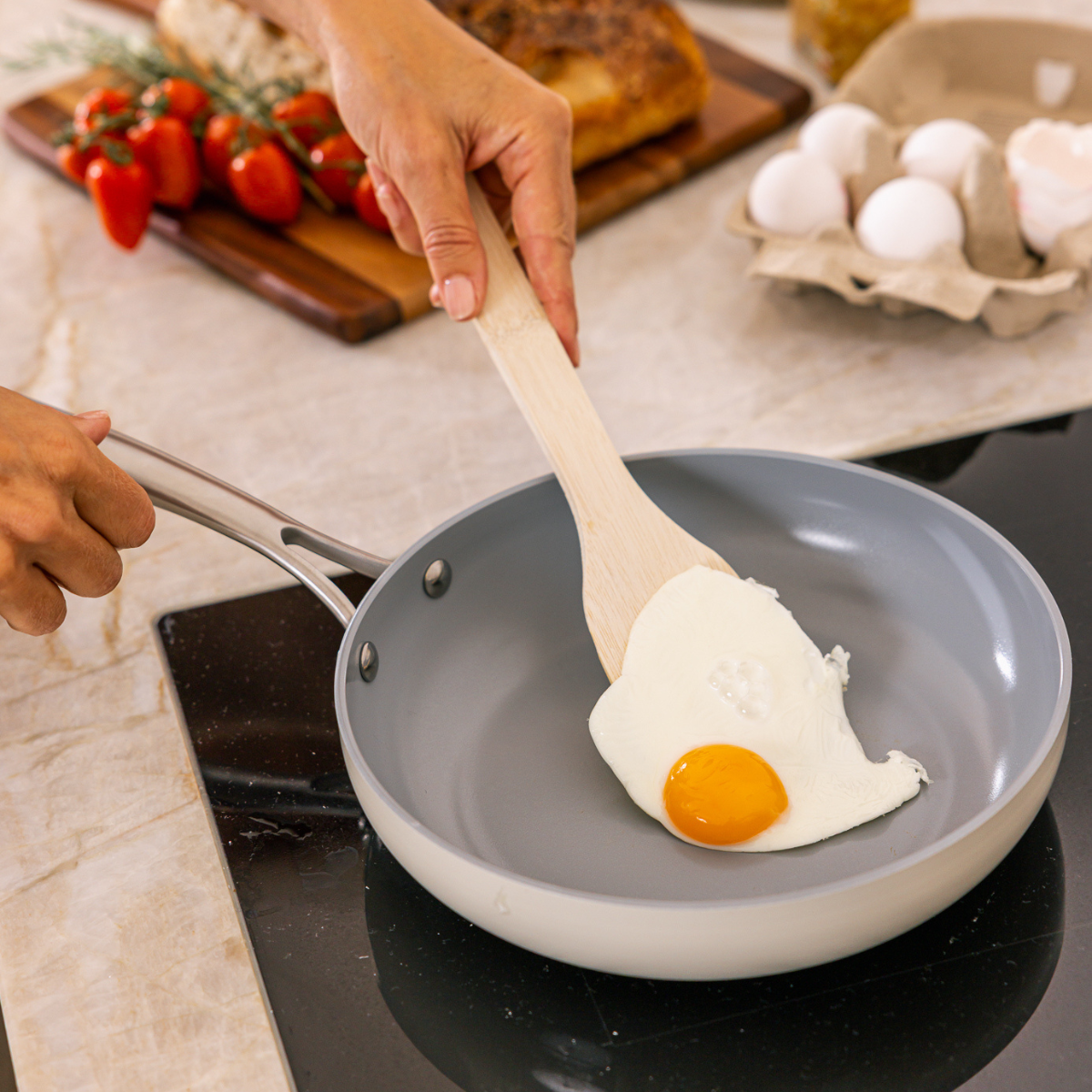 A hand flips a fried egg in a pan. Eggs, tomatoes, and bread are on the counter. The egg yolk is yellow, the white is white.