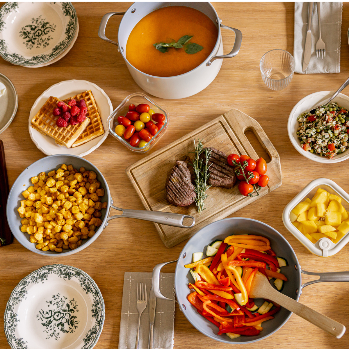 Wooden table with various dishes, including waffles, steak, vegetables, soup, and fruit, arranged for a meal.