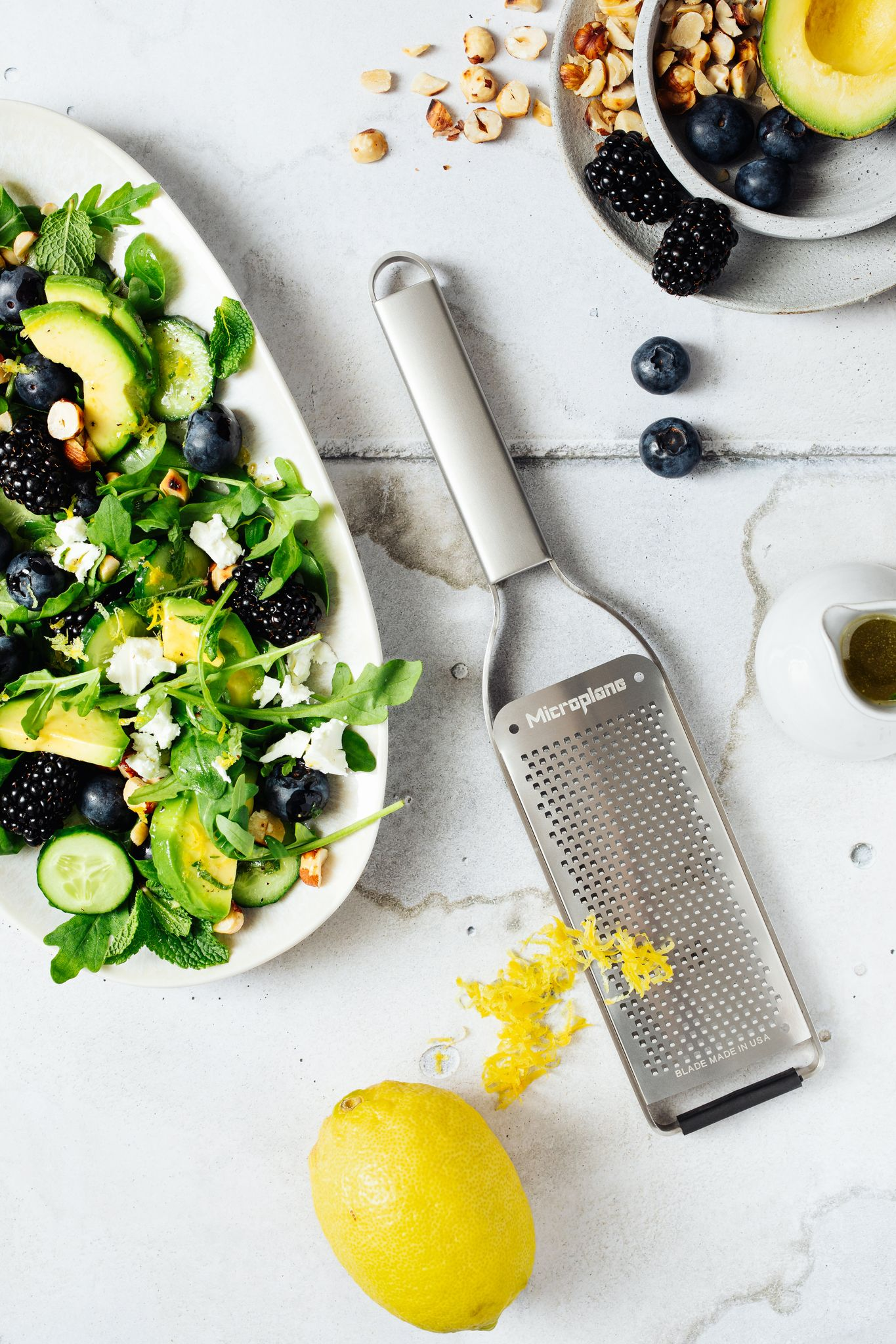 Ein lebendiger Salat mit Avocado, Blaubeeren, Brombeeren und Nüssen, plus Zitrone und Reibe auf weißer Oberfläche.