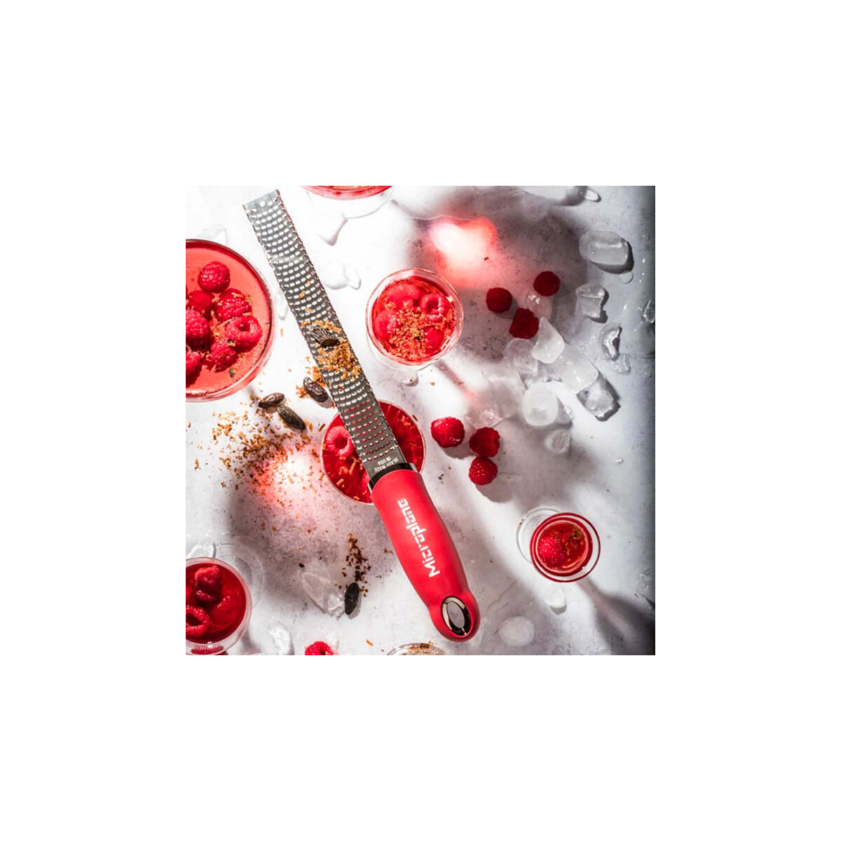 Ein roter Reibeisen mit Himbeeren und Getränken. Eiswürfel und verstreute Himbeeren sind ebenfalls auf einer weißen Oberfläche vorhanden.