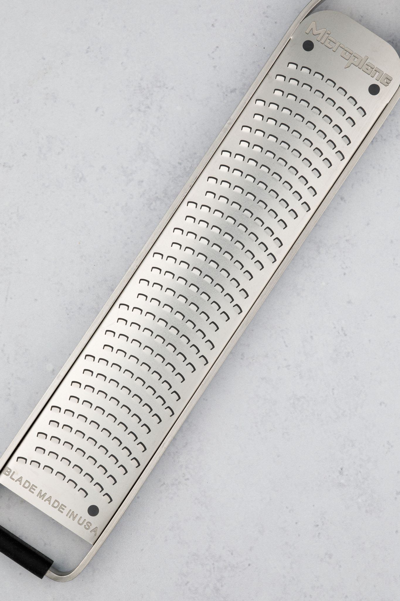 Ein Microplane-Reibeisen aus Edelstahl auf hellgrauer Oberfläche. Das Reibeisen ist rechteckig mit kleinen Klingen.