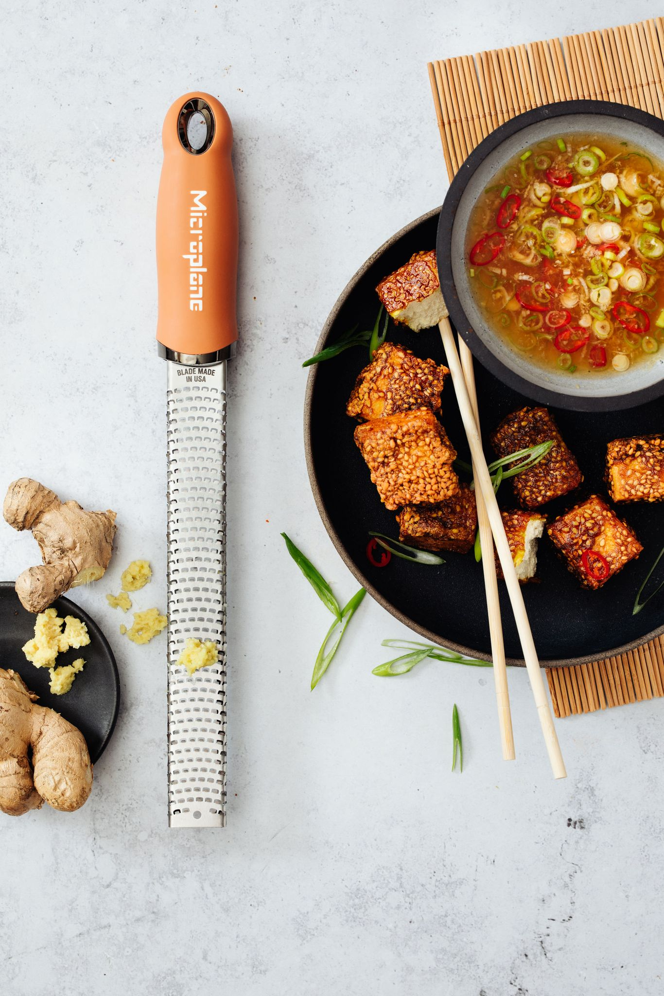 Reibe, Ingwer und Essen in einer Schüssel. Sesam-Tofu, Soße mit Frühlingszwiebeln und Chili sowie Essstäbchen auf Bambusmatte.