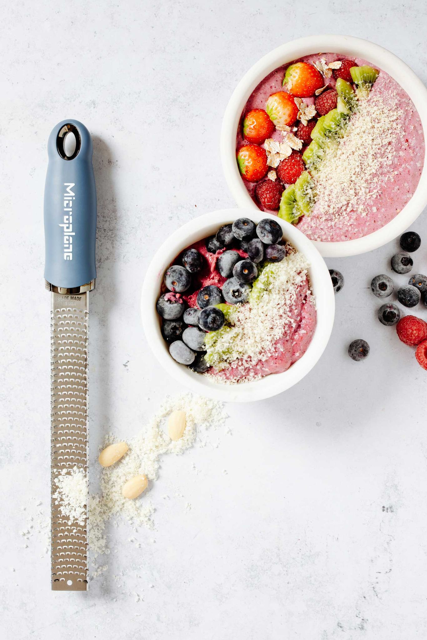 Zwei Smoothie-Schalen mit Beeren, Kiwi und geriebenen Mandeln. Eine Reibe und Mandeln sind daneben. Weißer Hintergrund.