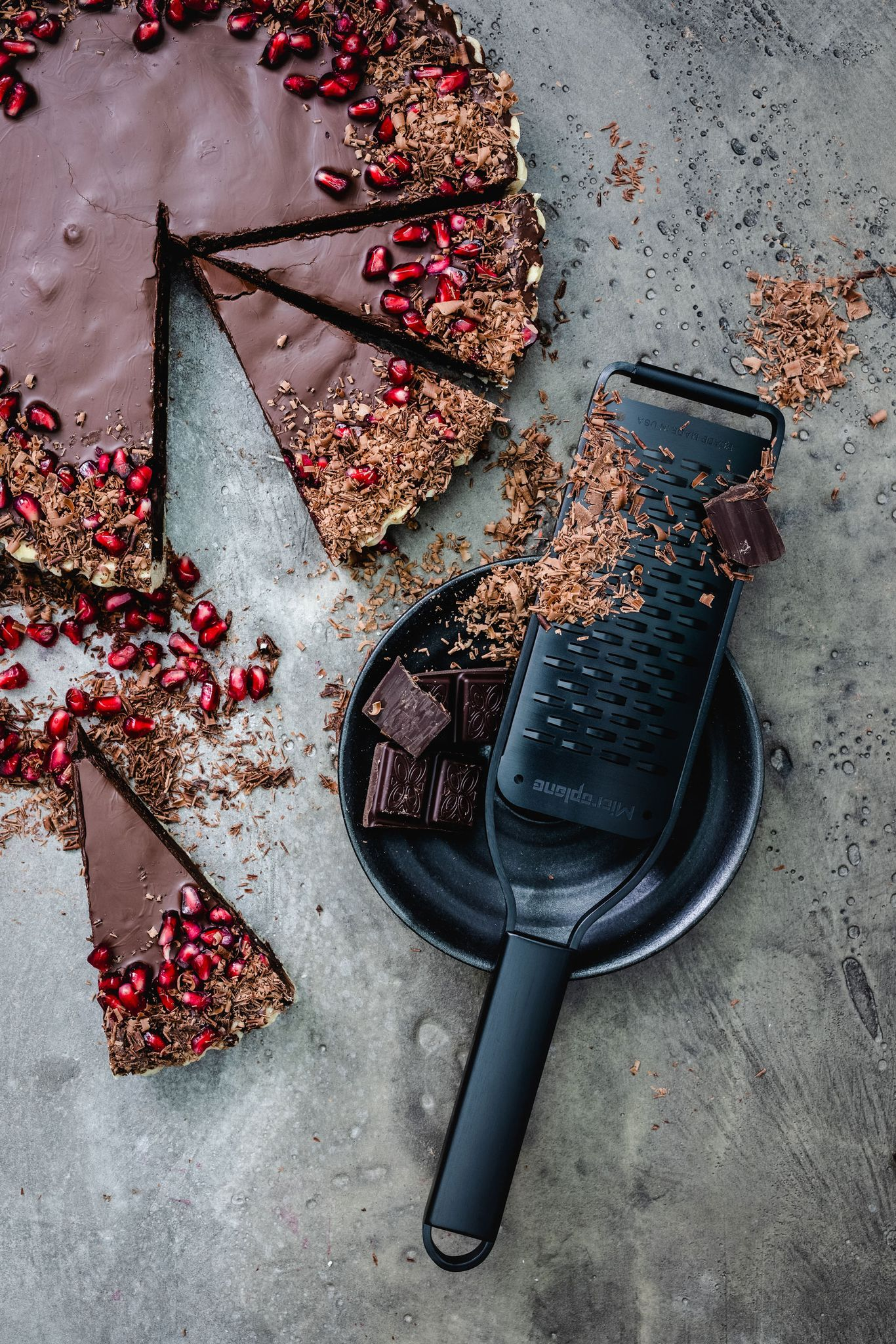 Ein Schokoladenkuchen mit Granatapfelkernen und Schokoladenraspeln, eine Reibe und Schokoladenstücke auf grauer Oberfläche.